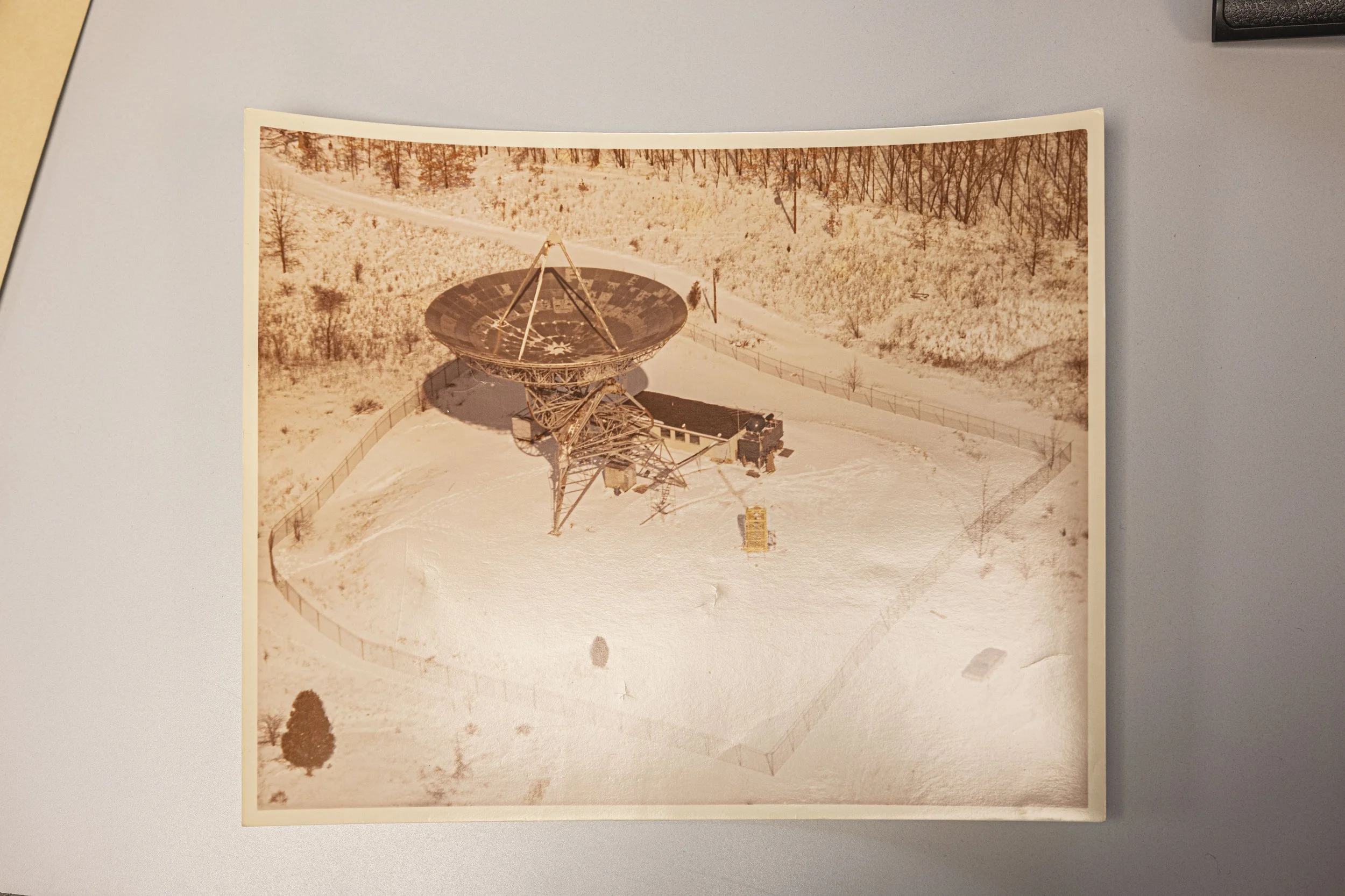 A large radio telescope in a snow-covered landscape surrounded by a fence and sparse trees.