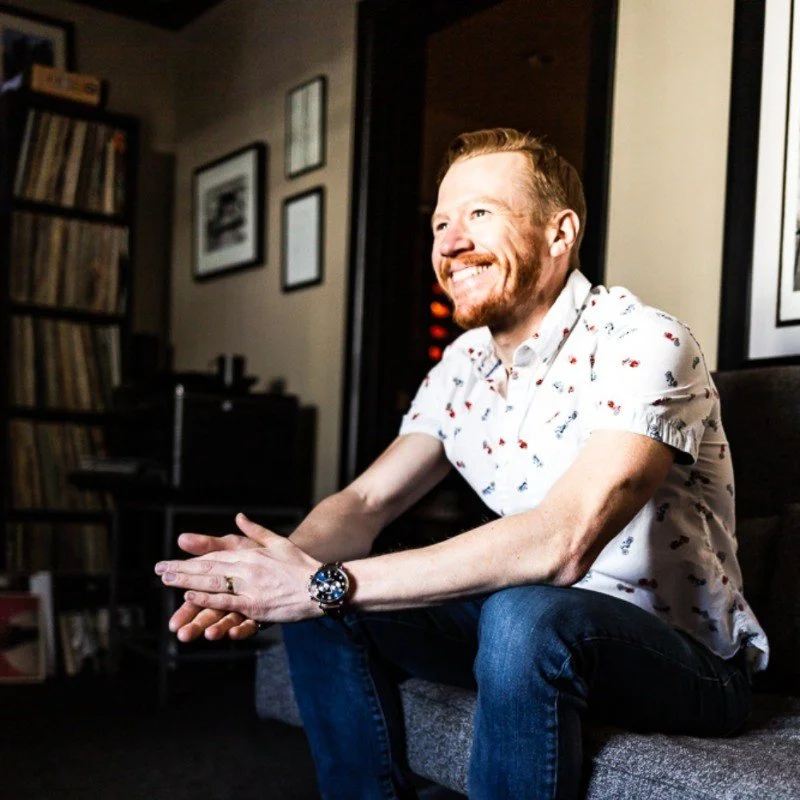 A smiling man with a beard and short hair sitting on a gray sofa in a room decorated with framed pictures and a bookshelf, wearing a white shirt with a small pattern, jeans, and a wristwatch.