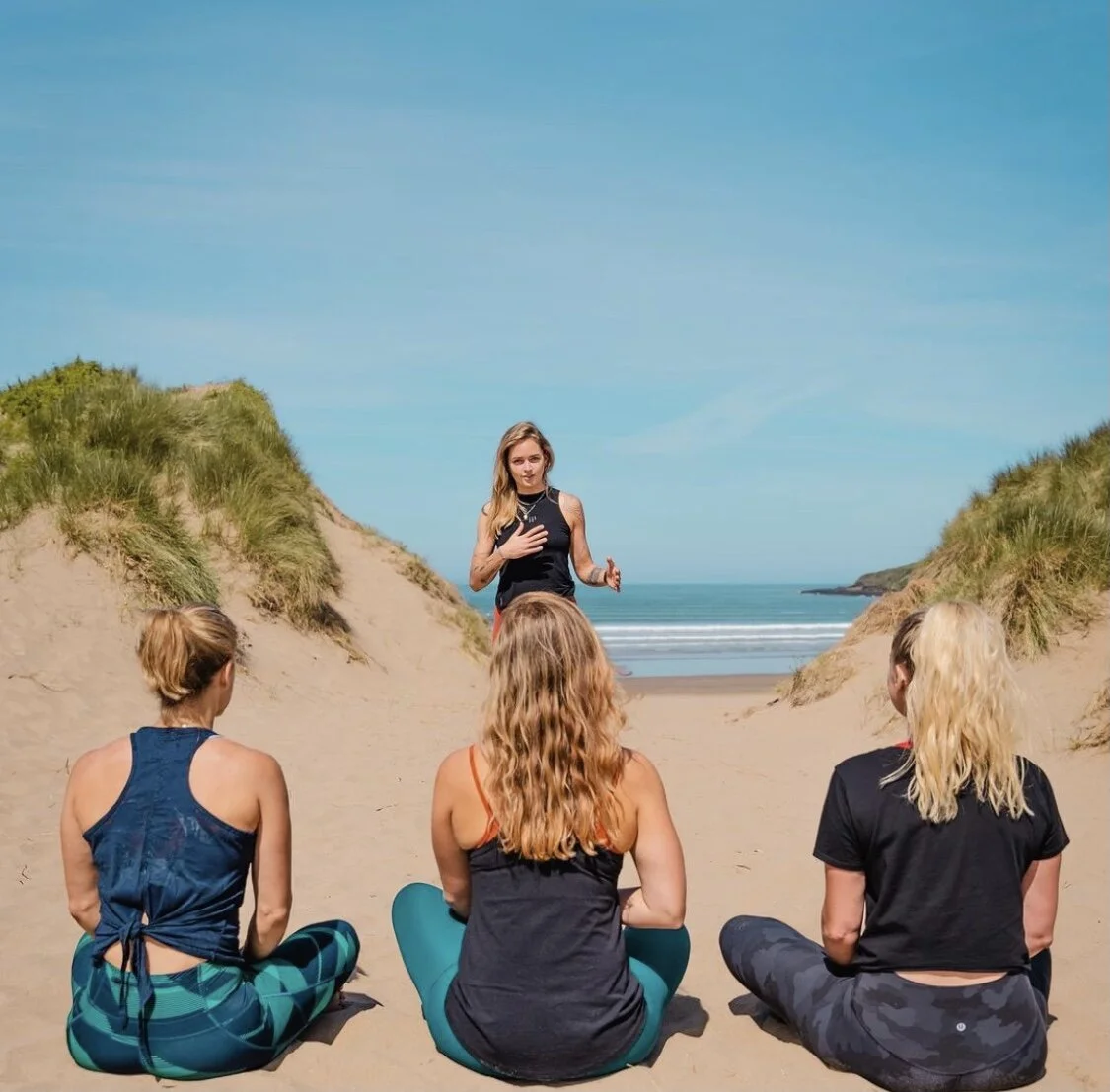 A woman giving a yoga or meditation class on the beach with three women sitting on the sand, facing her and listening.