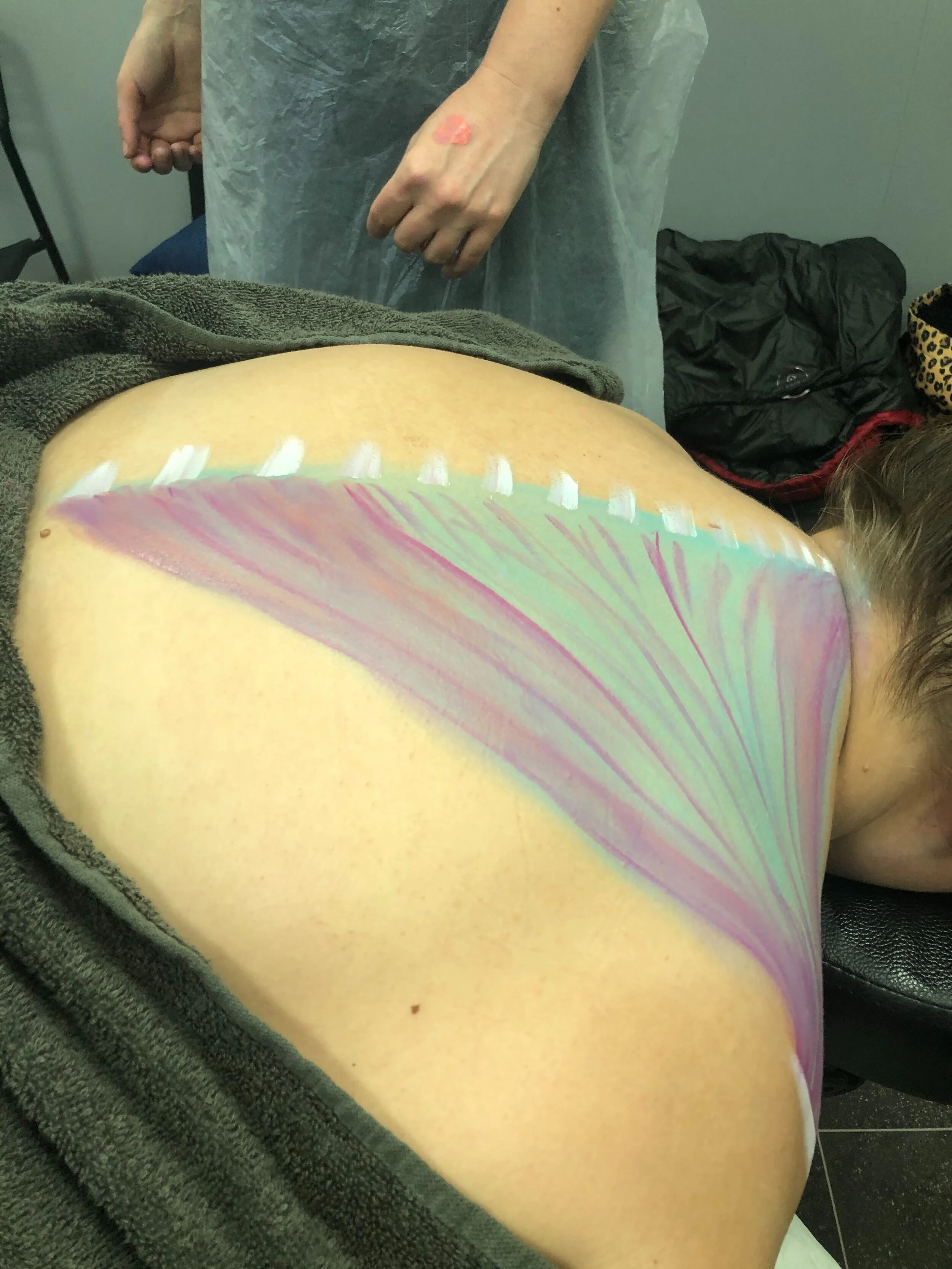 Someone lying face down with colorful butterfly wing body art on their back, while a person standing nearby is in a medical gown in a clinical setting.