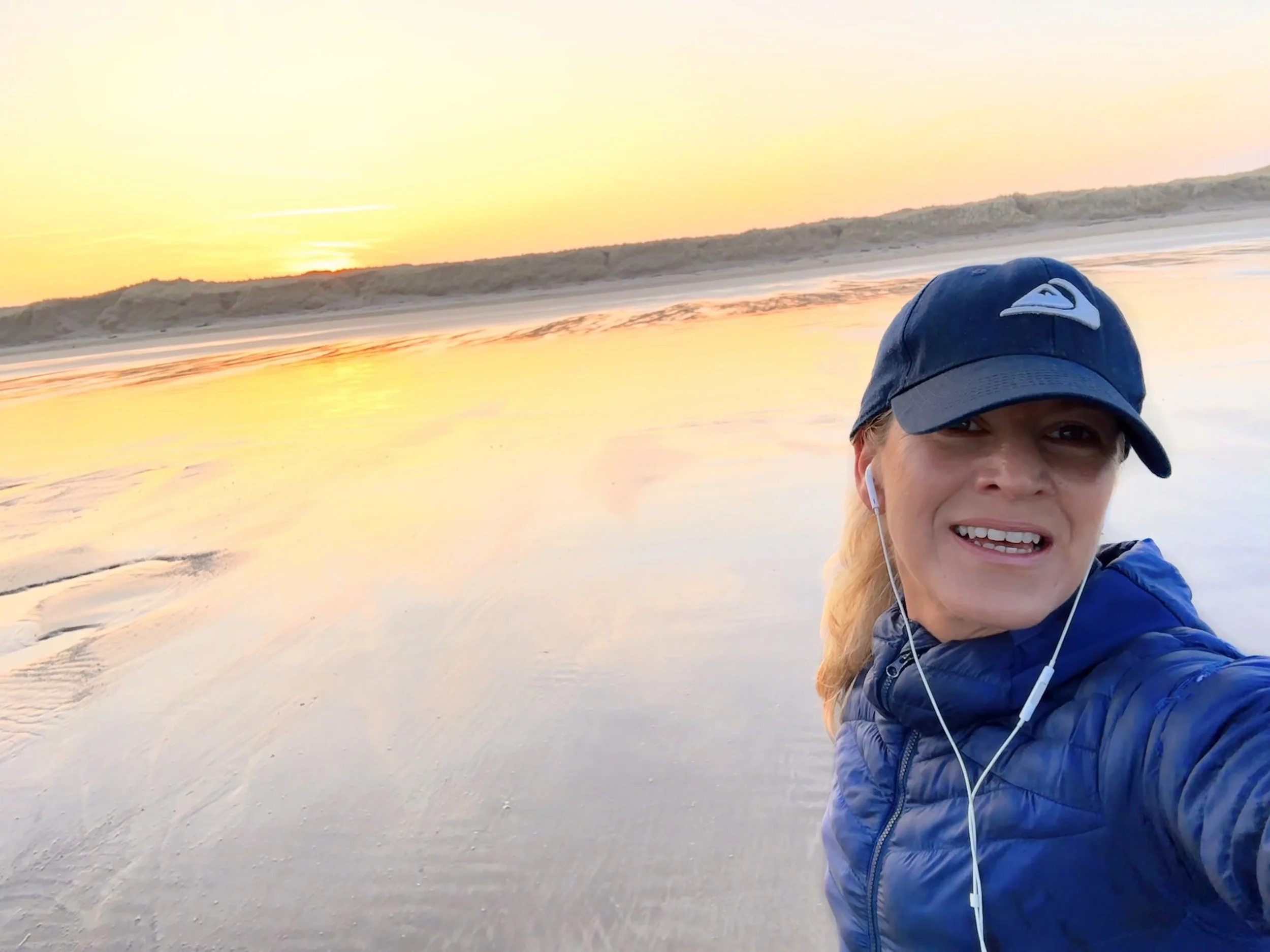 A woman taking a selfie on a beach at sunrise, wearing a blue jacket, cap, and earphones, with the wet sand and ocean behind her.