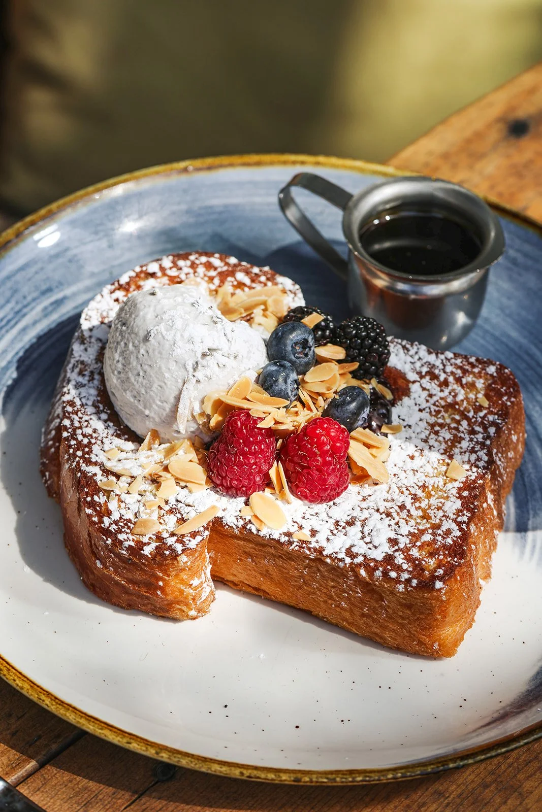 A slice of French toast topped with powdered sugar, fresh berries, whipped cream, and almond slices, served with a small pitcher of syrup.