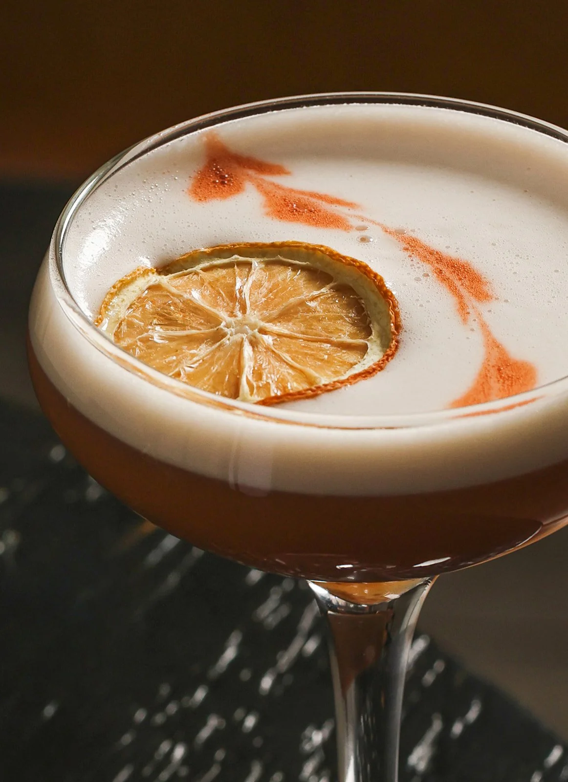 Close-up of a cocktail with a lemon slice and a frothy white top, in a coupe glass.