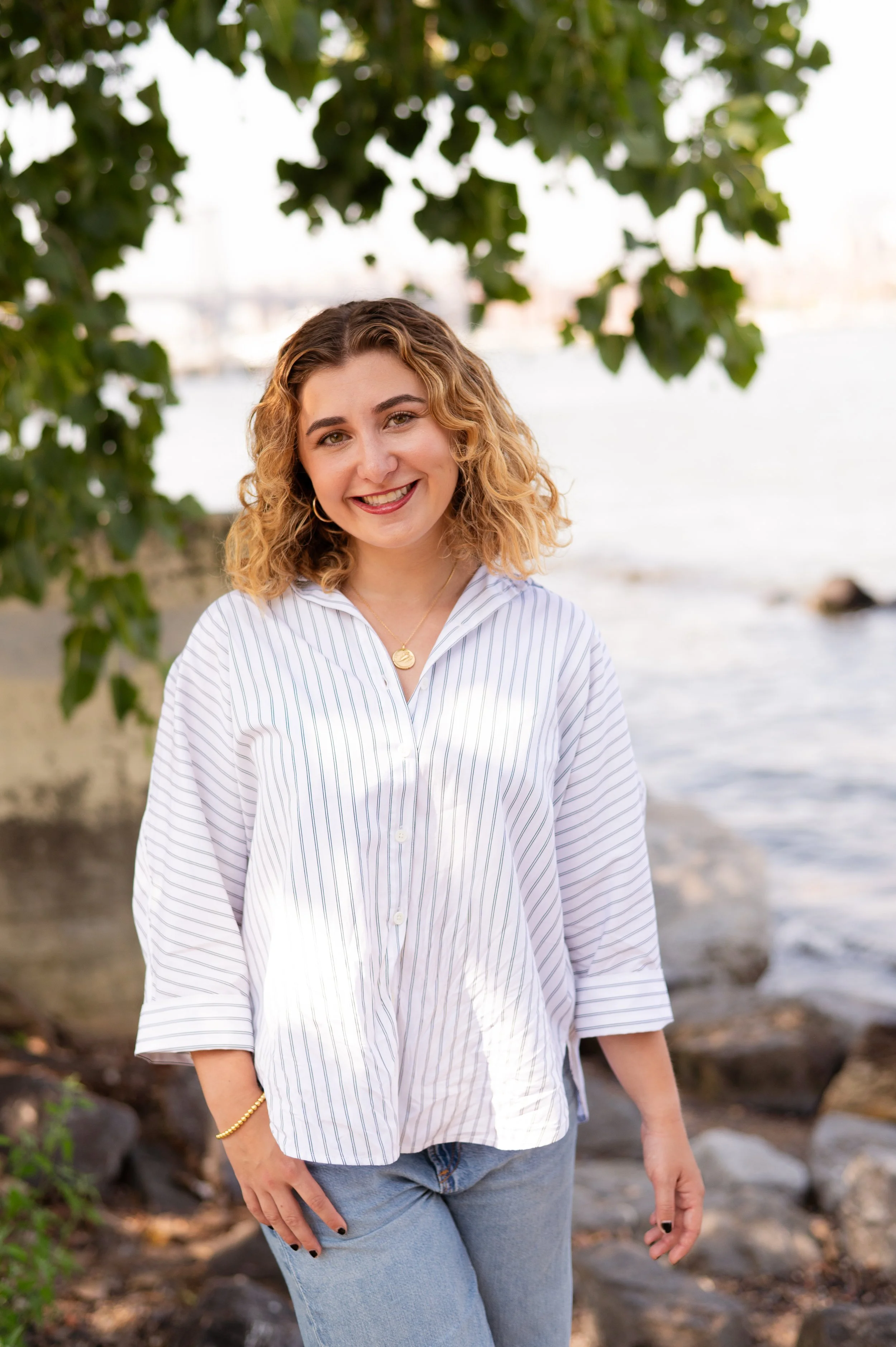 A woman outside in front of water and rocks and trees. She has curly blonde hair.  She is wearing a striped long-sleeve shirt.