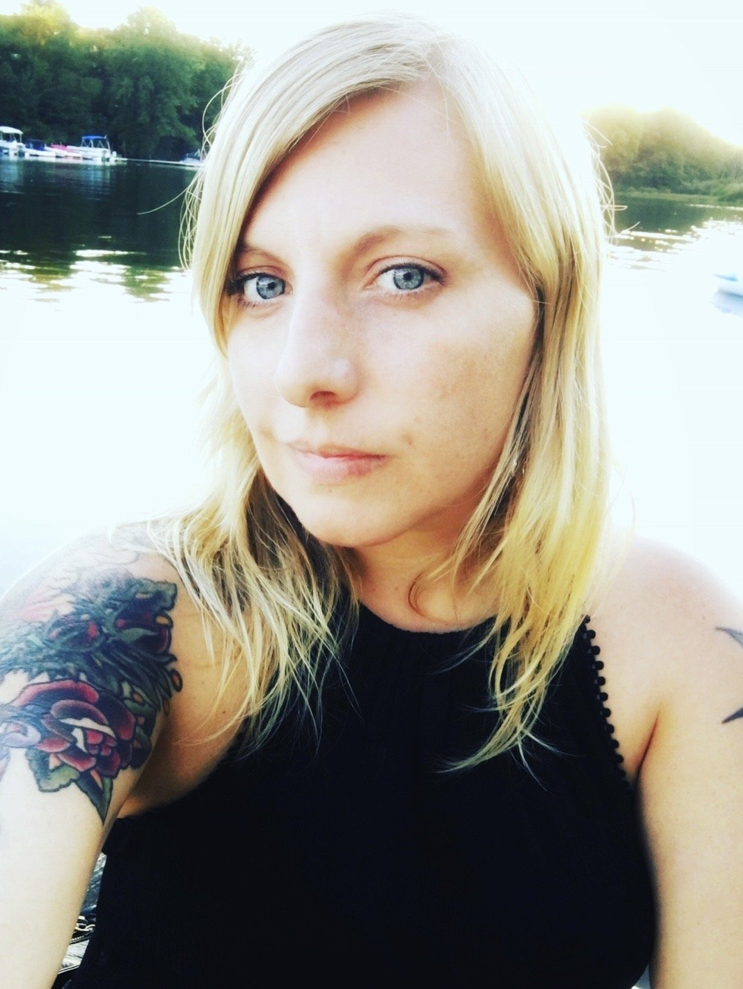 Woman infront of water. She has blonde shoulder length hair, tucked behind her ears. She has blue eyes.