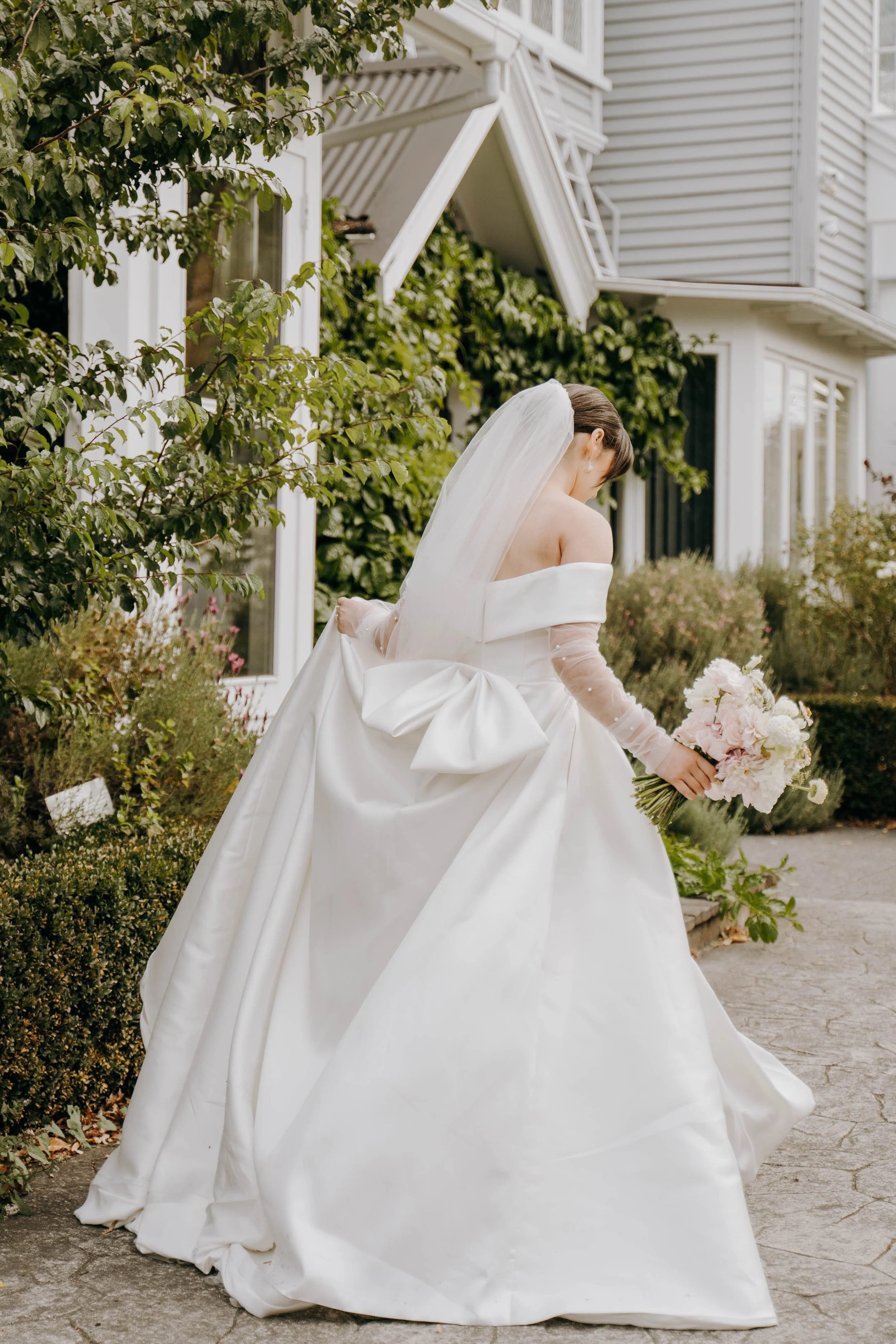 Bride in a white wedding gown holding a bouquet of pink and white flowers, walking on a sidewalk next to a garden with green bushes and a corner of a white house in the background.