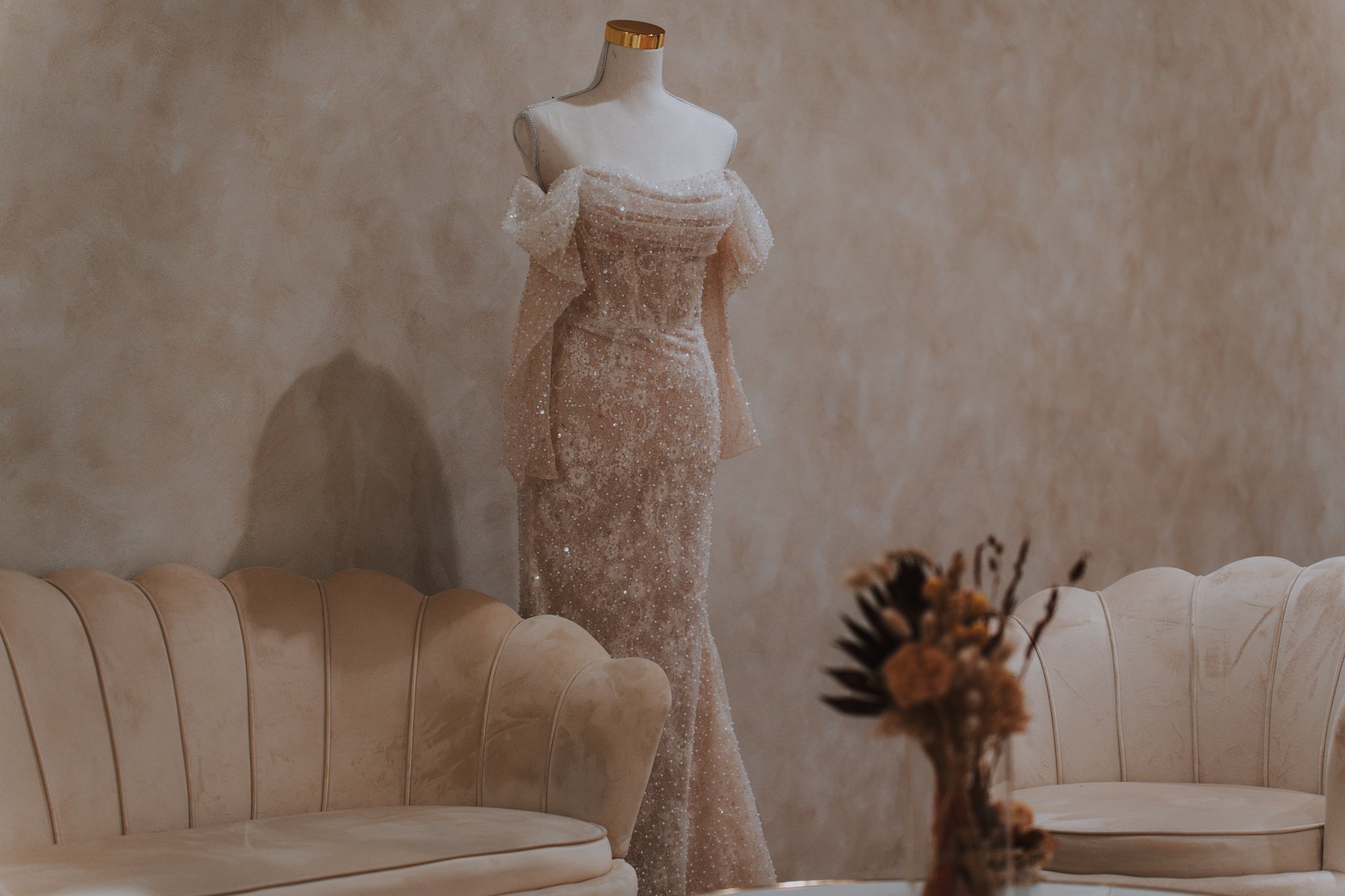 Elegant, sparkle-embellished off-the-shoulder gown displayed on a mannequin in a softly lit room with vintage-style beige sofas and a blurred bouquet in the foreground.