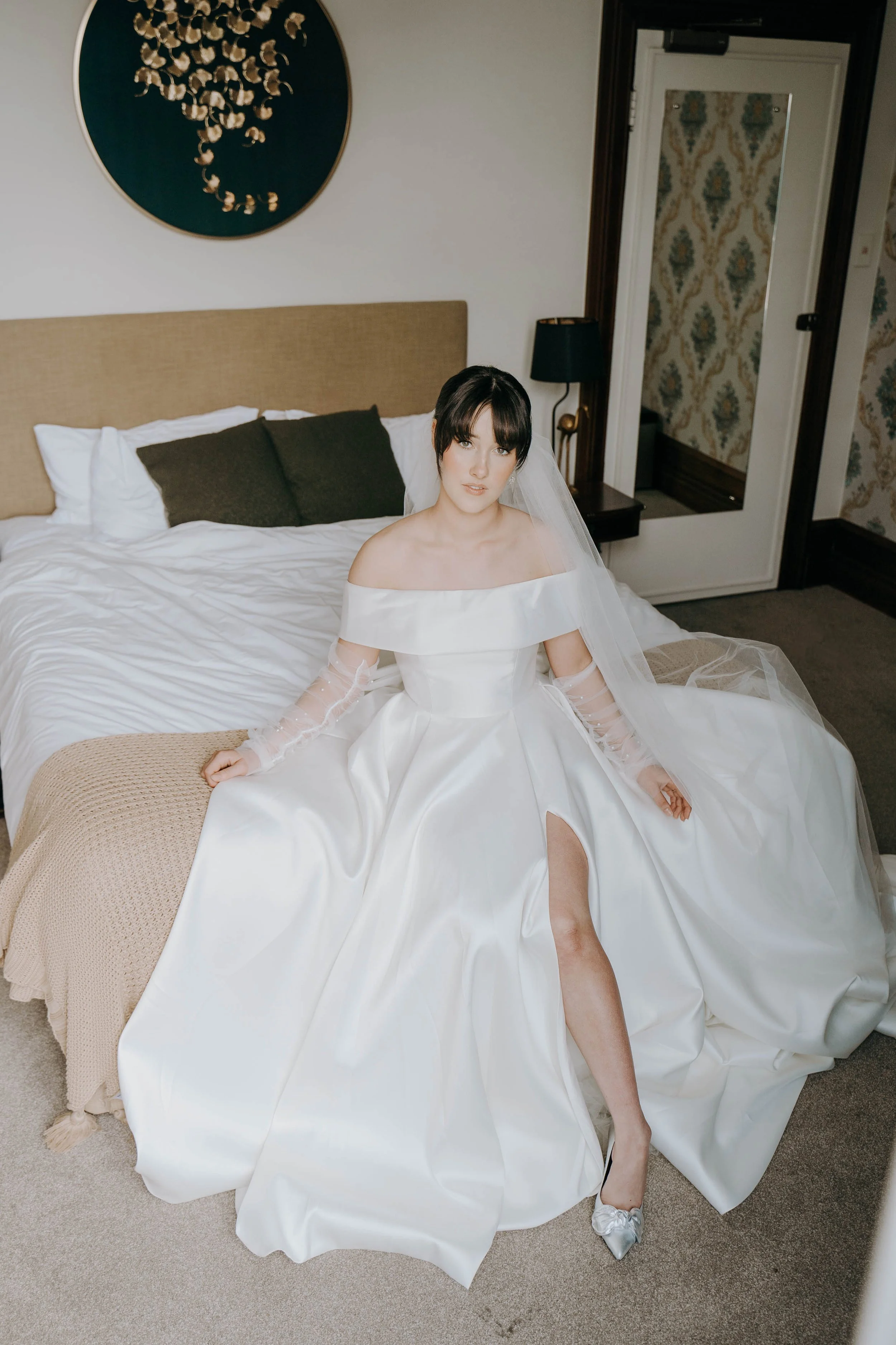 A bride in a white wedding gown sitting on the edge of a bed in a hotel room, with a slit showing her right leg. She has dark hair with bangs and is wearing a veil.