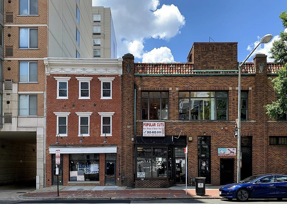 Street view of three adjacent brick buildings, one with a barber shop and a sign for popular cuts, another with windows and a storefront.