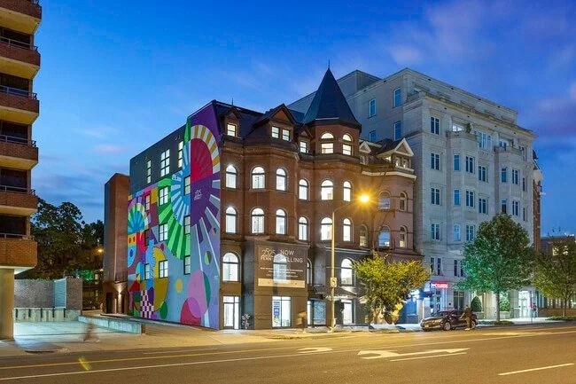 City street scene featuring a colorful, multi-story building with mural art, next to a white building and trees at dusk.