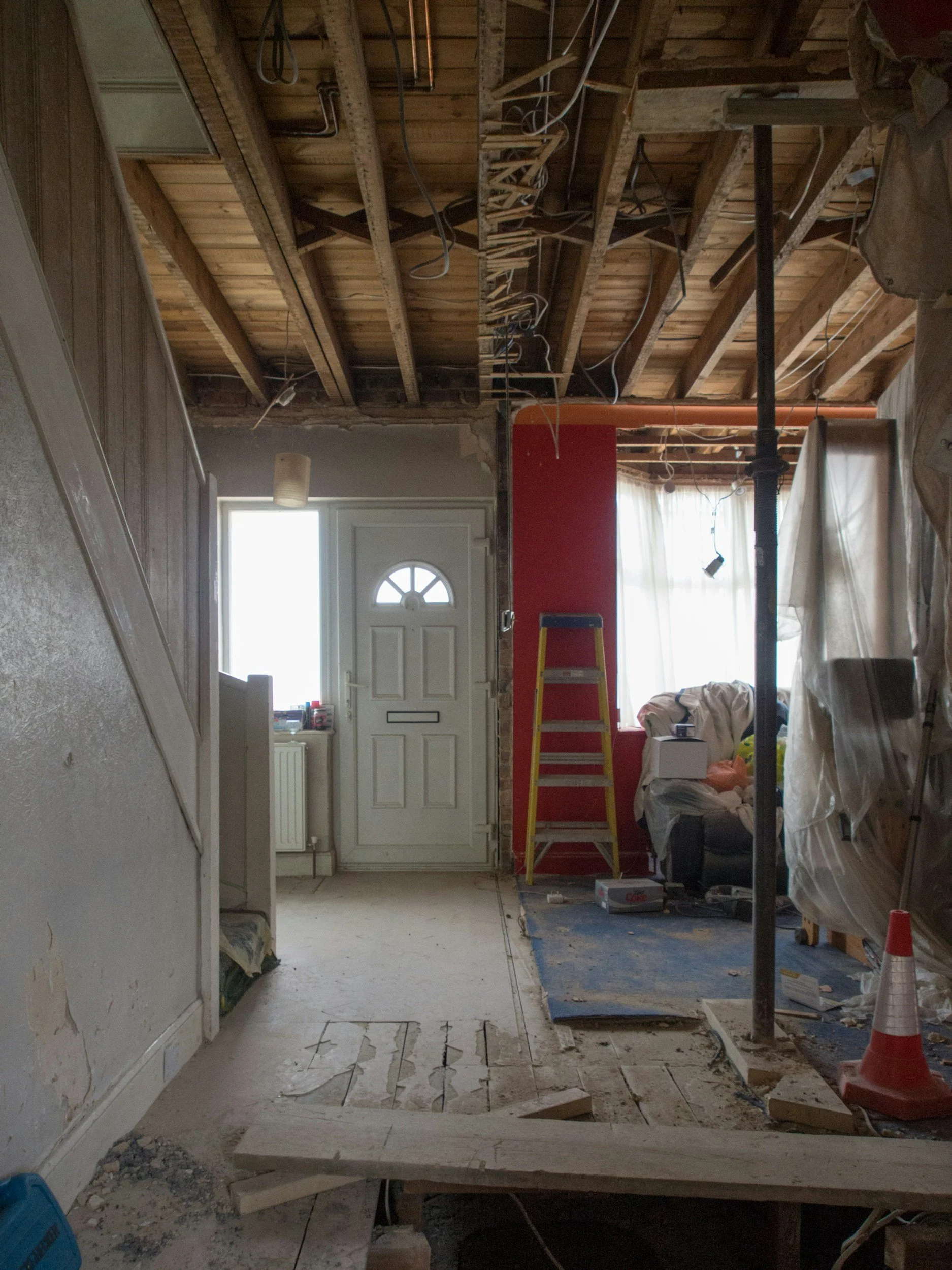 Interior of a house under renovation with exposed ceiling joists, hanging electrical wires, a partially removed ceiling, and construction materials scattered on the floor.