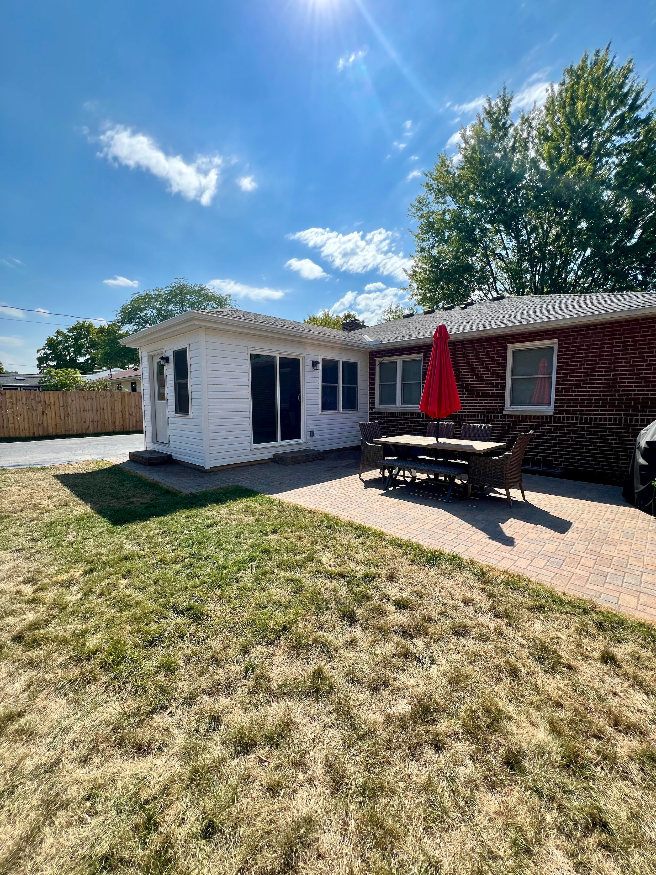 A backyard with a brick and white siding house featuring a small patio with a table and chairs, a red umbrella, and an uneven grassy lawn under a bright sunny sky with few clouds.