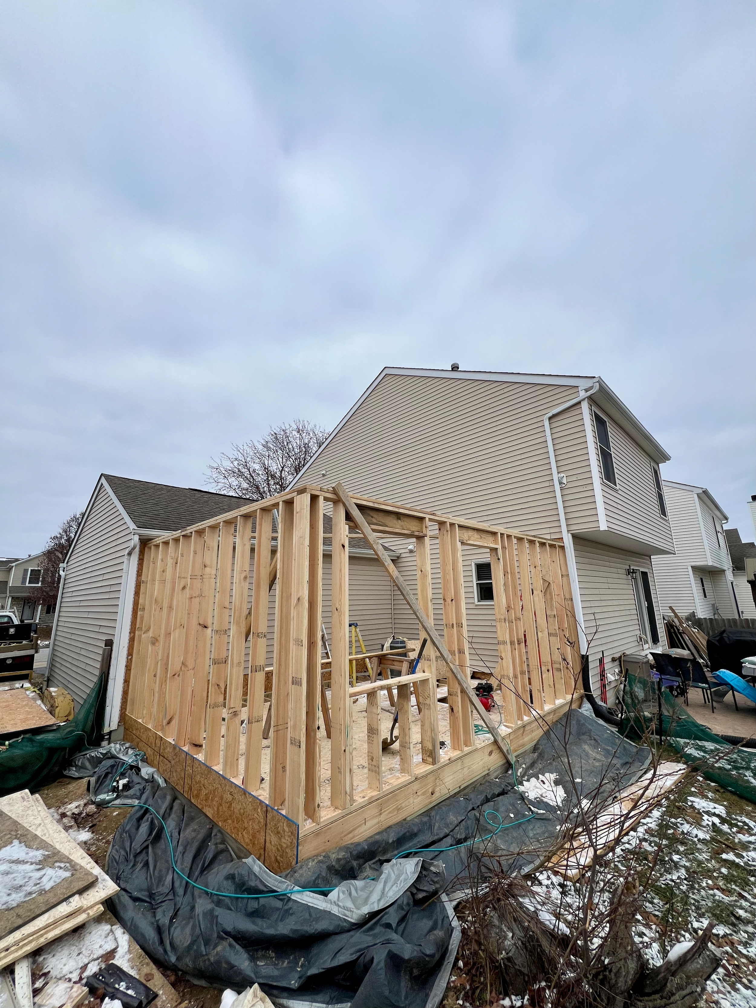 A house under construction with a wooden frame for an addition or extension, with other houses visible in the background on a cloudy day.