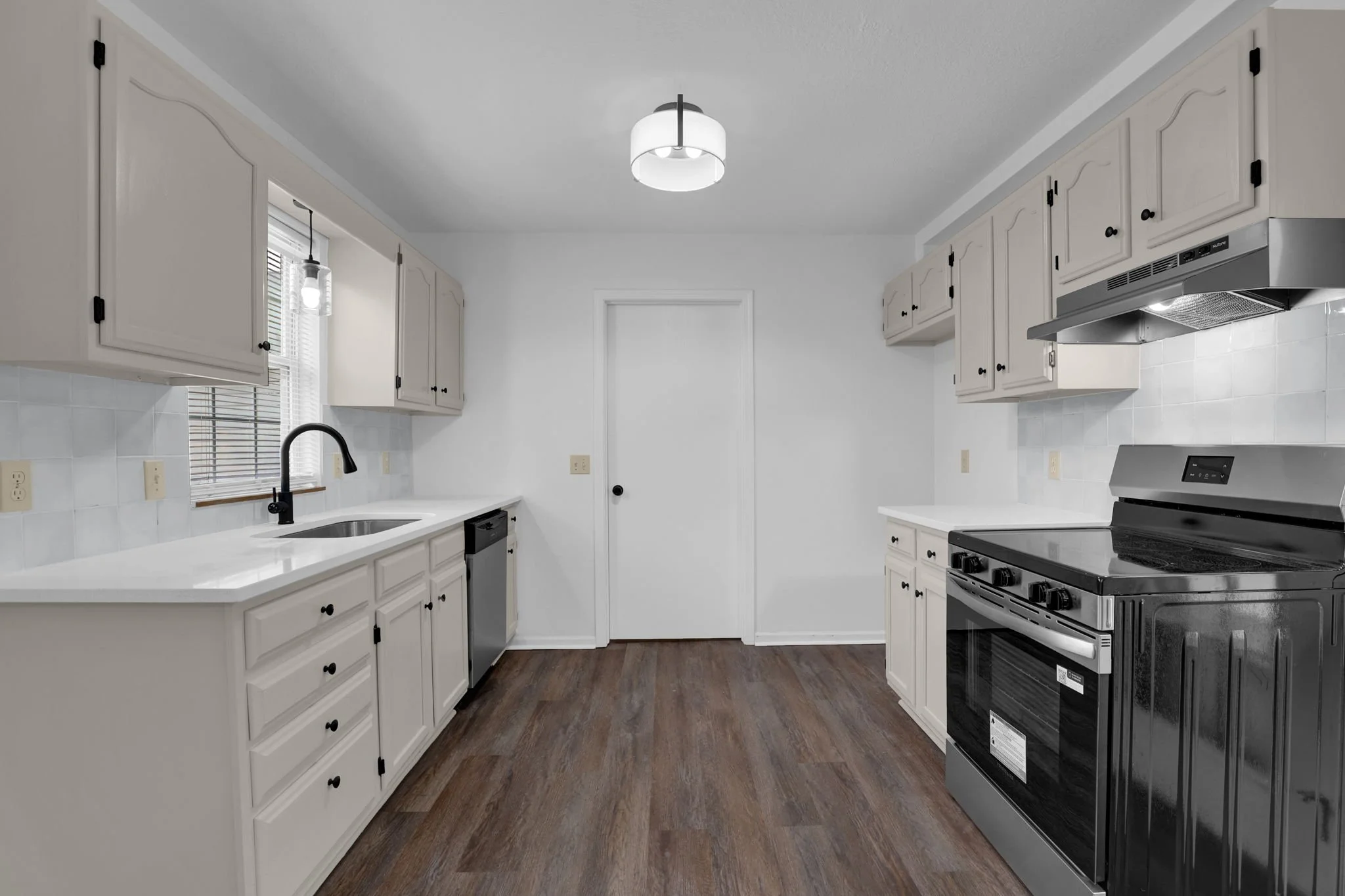A bright white kitchen with white cabinets, black hardware, a black faucet, a stainless steel stove, and white countertops. There is a window with blinds on the left and a ceiling light fixture.
