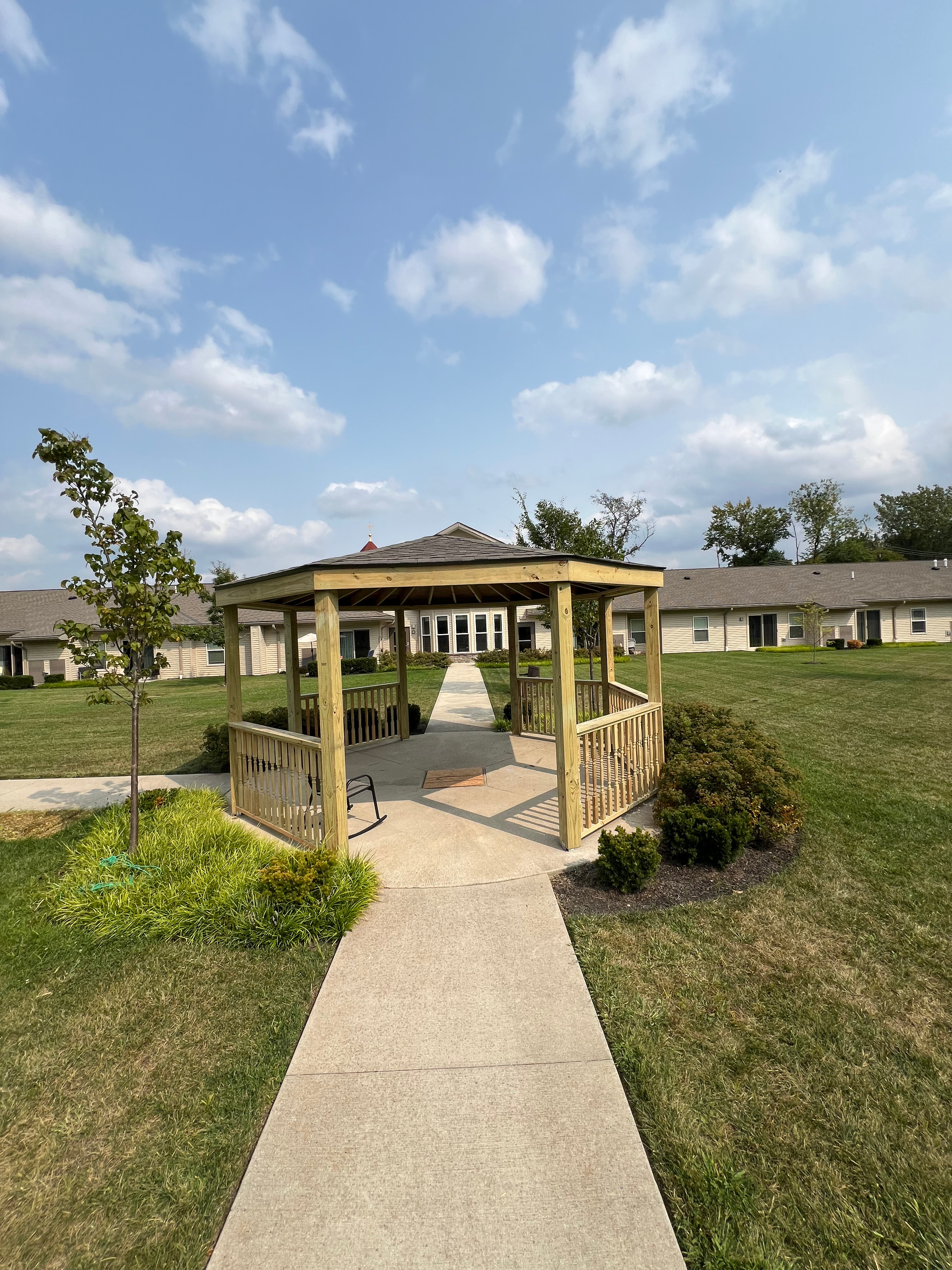 A sidewalk leading to a small wooden gazebo in a grassy outdoor area, with apartment buildings in the background under a partly cloudy sky.
