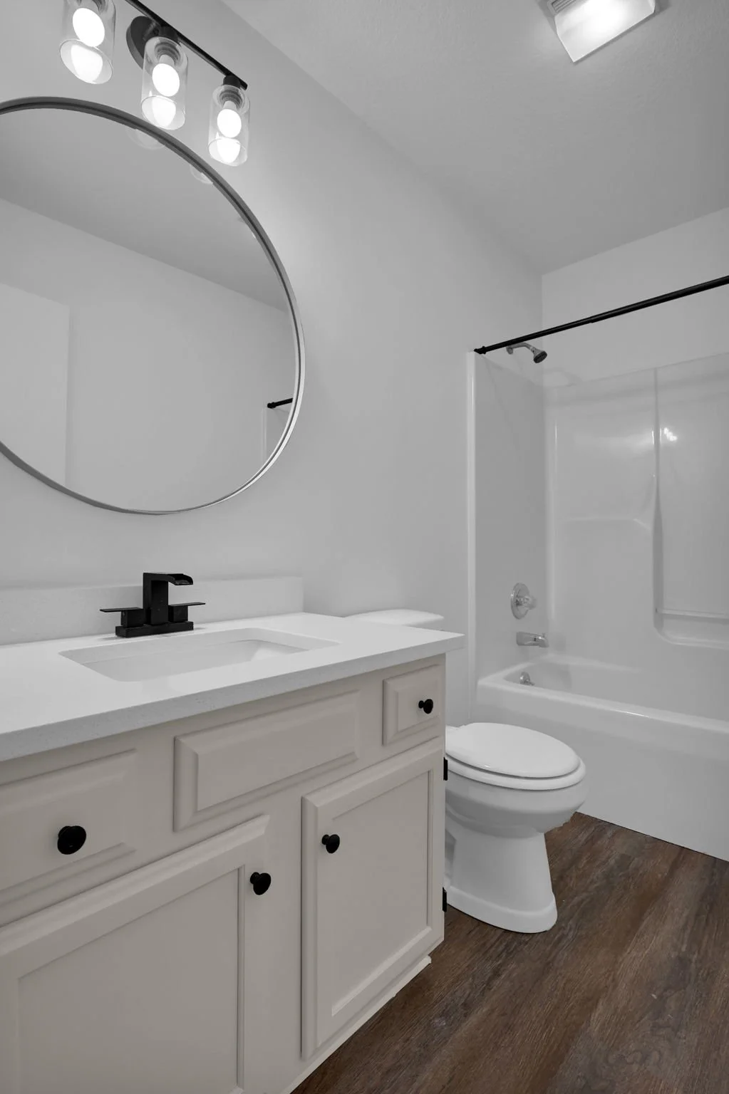 Modern bathroom with white vanity, black faucet, round mirror, toilet, bathtub with shower, wood-look flooring, and ceiling light fixture.
