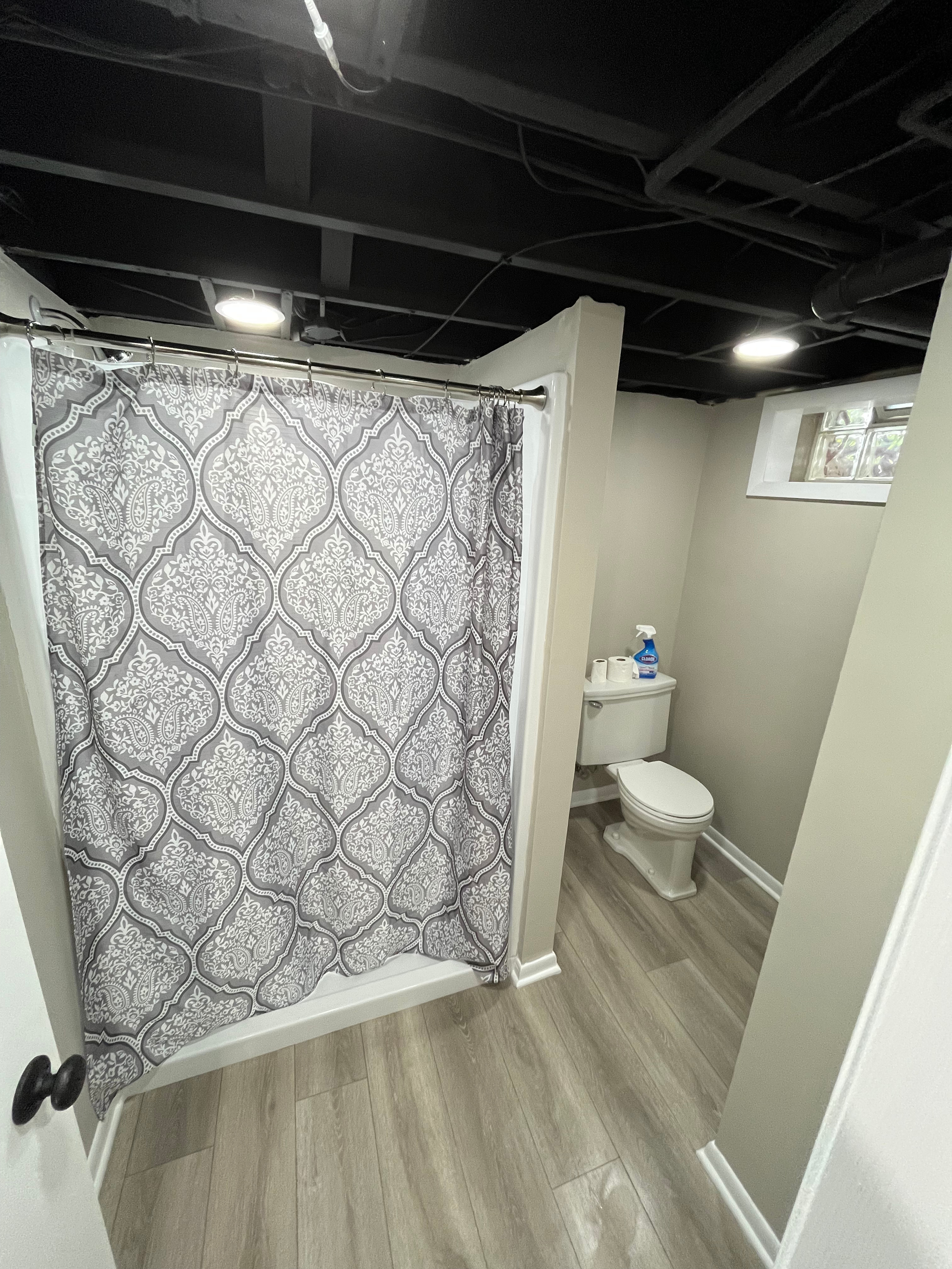 A small bathroom with a shower curtain, a toilet, and a window. The floor is wood-look vinyl, and ceiling has exposed black pipes and beams.