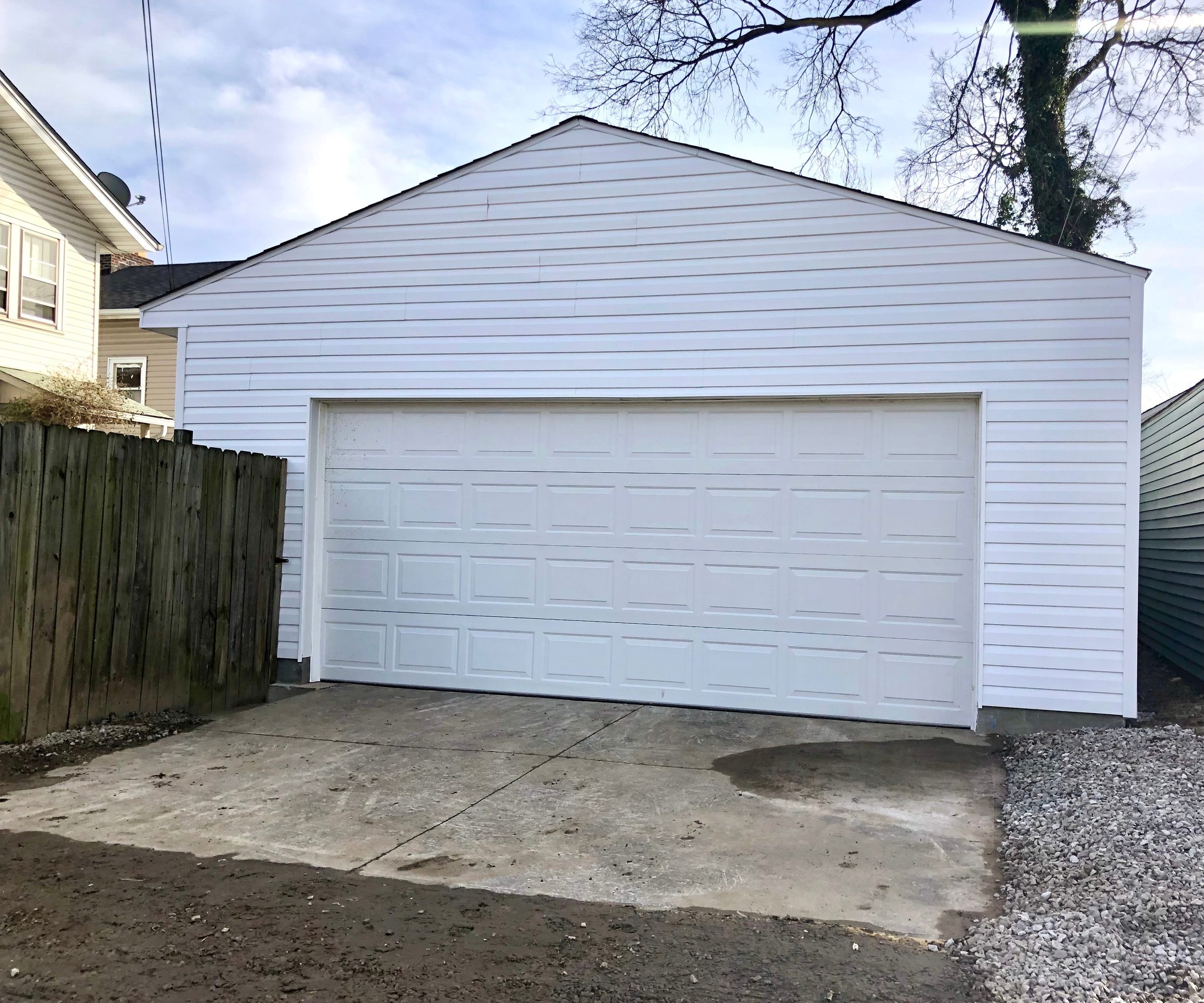 A newly built white garage with a closed garage door, adjacent to a wooden fence and neighboring houses, under a partly cloudy sky.