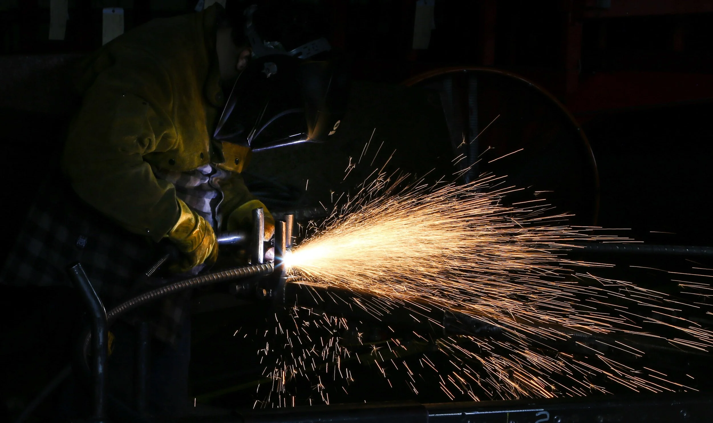 A worker in protective gear welding metal, producing sparks in a dark setting.