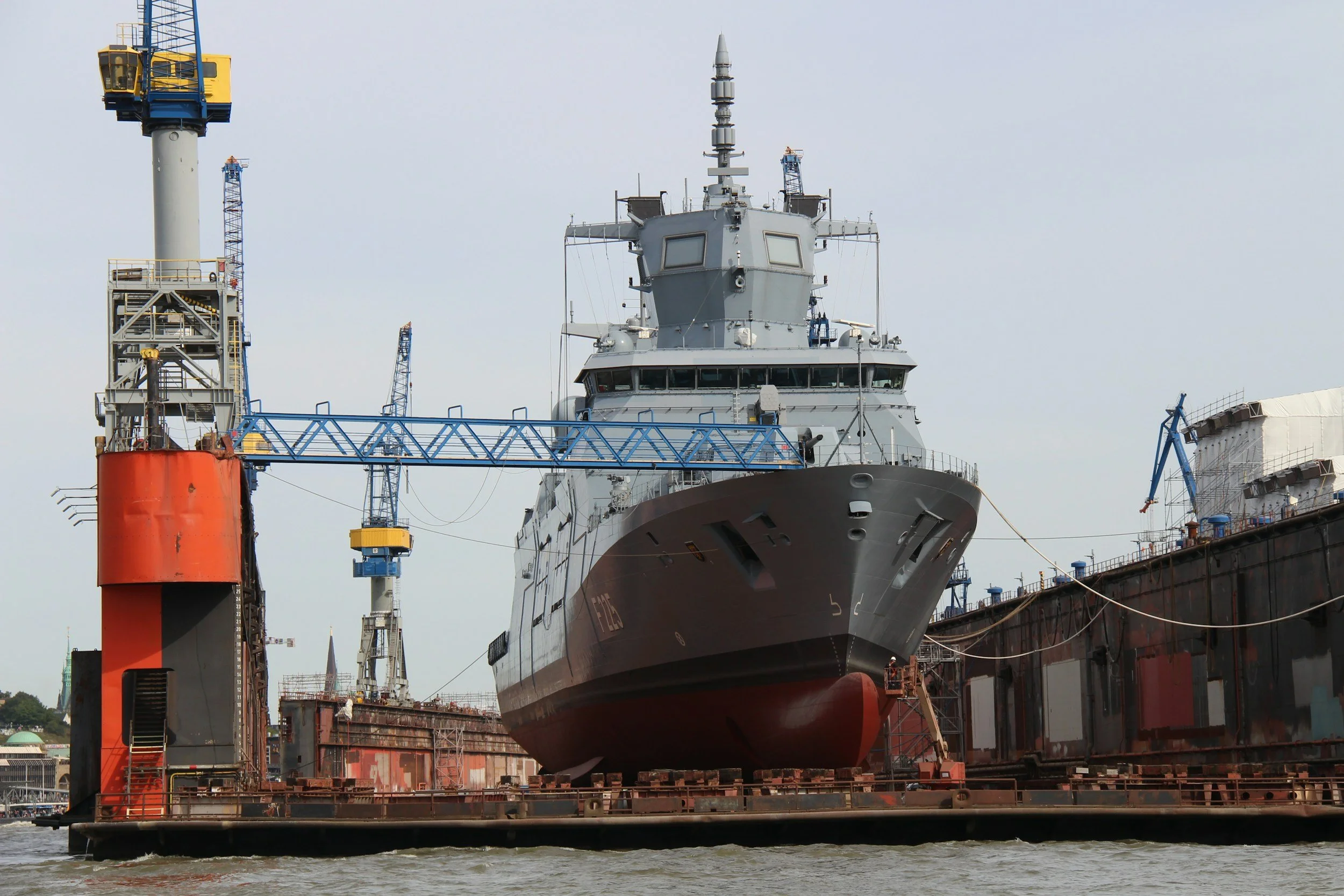 A large naval ship docked in a shipyard with surrounding scaffolding and cranes, under an overcast sky.