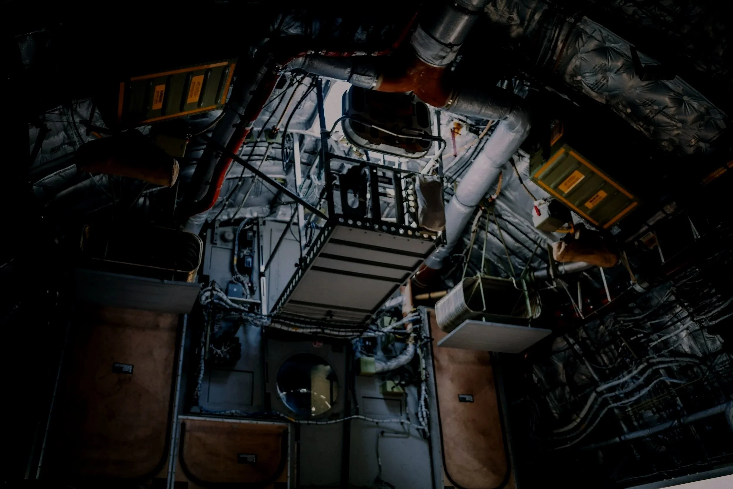 Interior view of a spacecraft or space station module with various pipes, cables, and equipment, viewed from below through a hatch or opening.