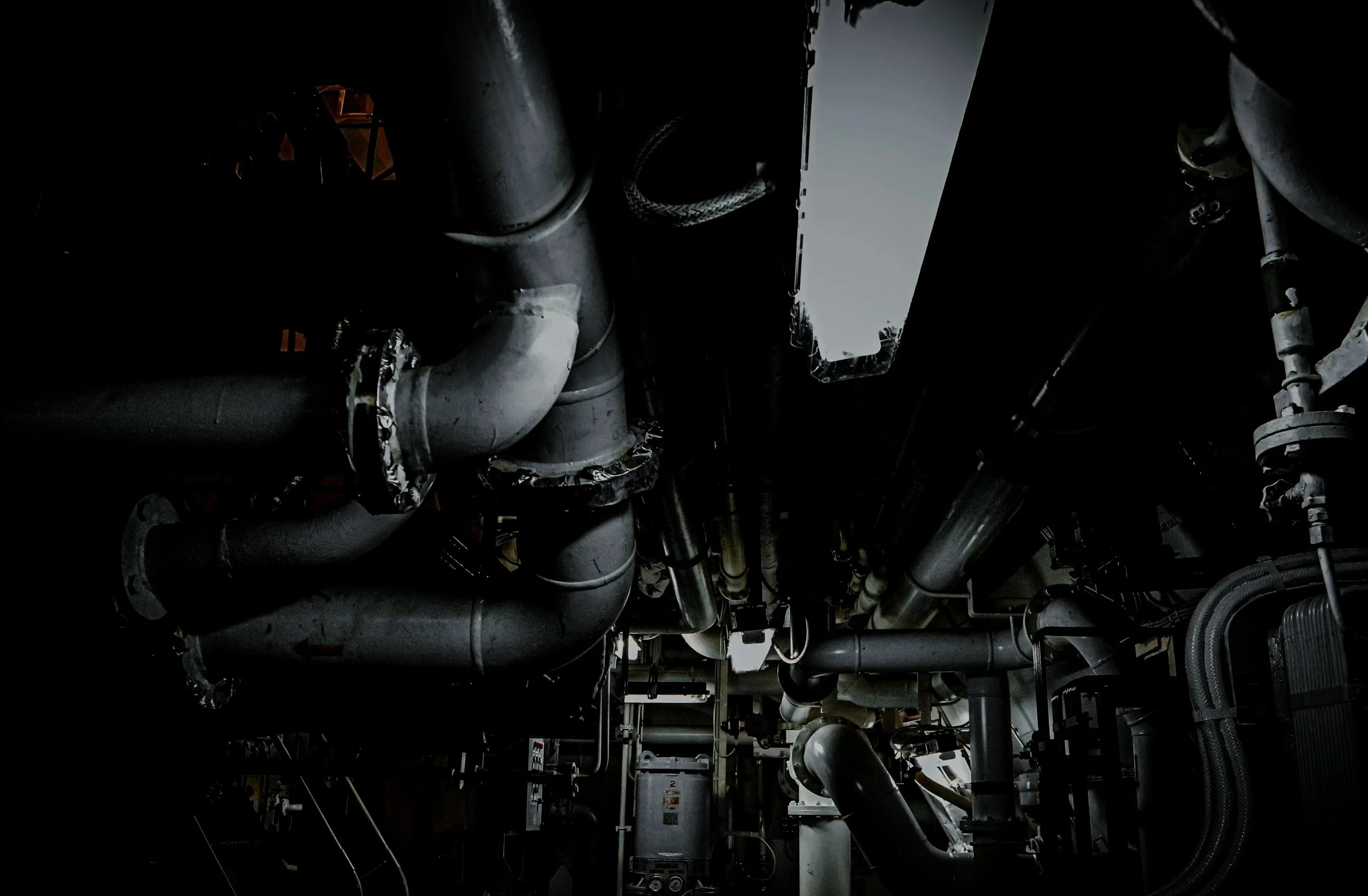 Dark industrial scene with large pipes and machinery, including valves, gauges, and electrical panels.