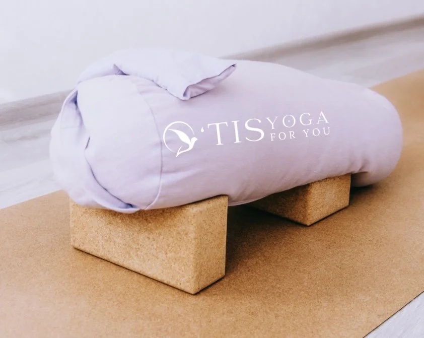 A yoga studio with a view of a large body of water through three windows, decorated with statues and ornaments, and a colorful yoga mat on the wooden floor.