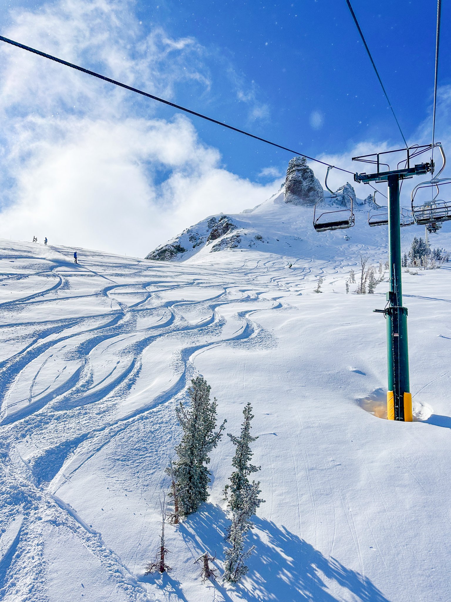 Snow-covered mountain landscape with ski lift chairs and multiple ski tracks in the snow, under a partly cloudy blue sky.