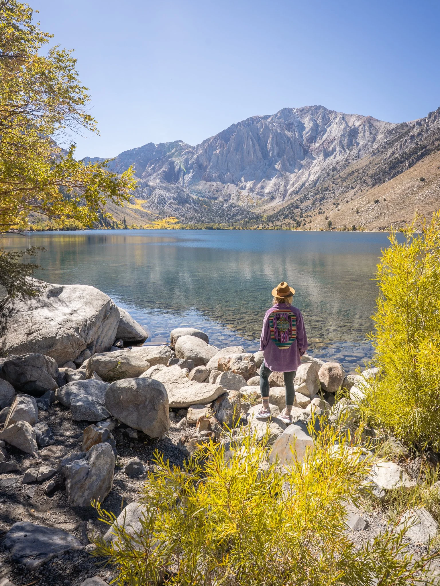 A woman standing on rocks by a lake, looking at mountains with snow patches under a clear blue sky, surrounded by green and yellow foliage.