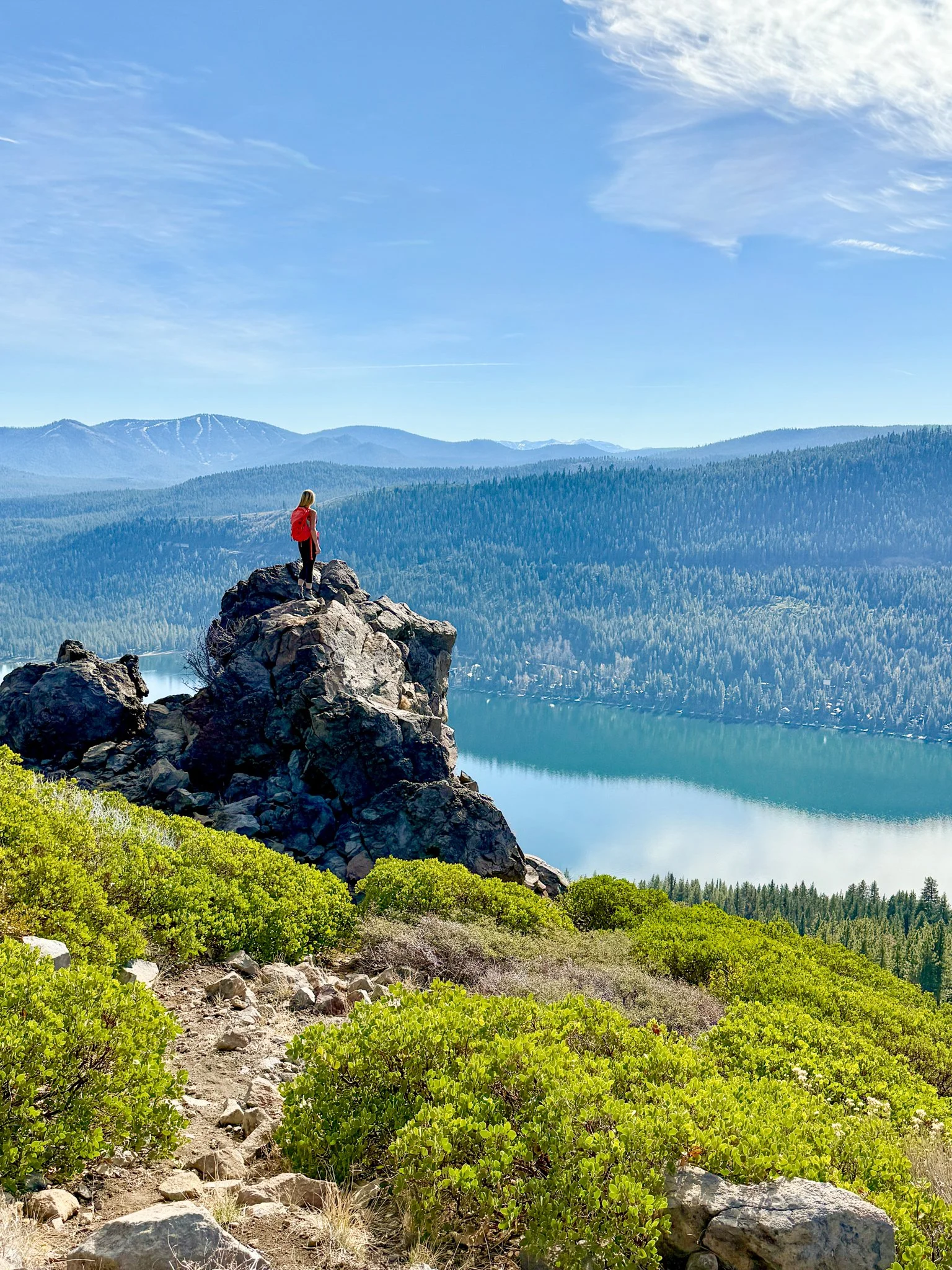 A hiker in a red backpack stands on a rocky outcrop overlooking a large body of water, surrounded by green hills and mountains under a blue sky with wispy clouds.