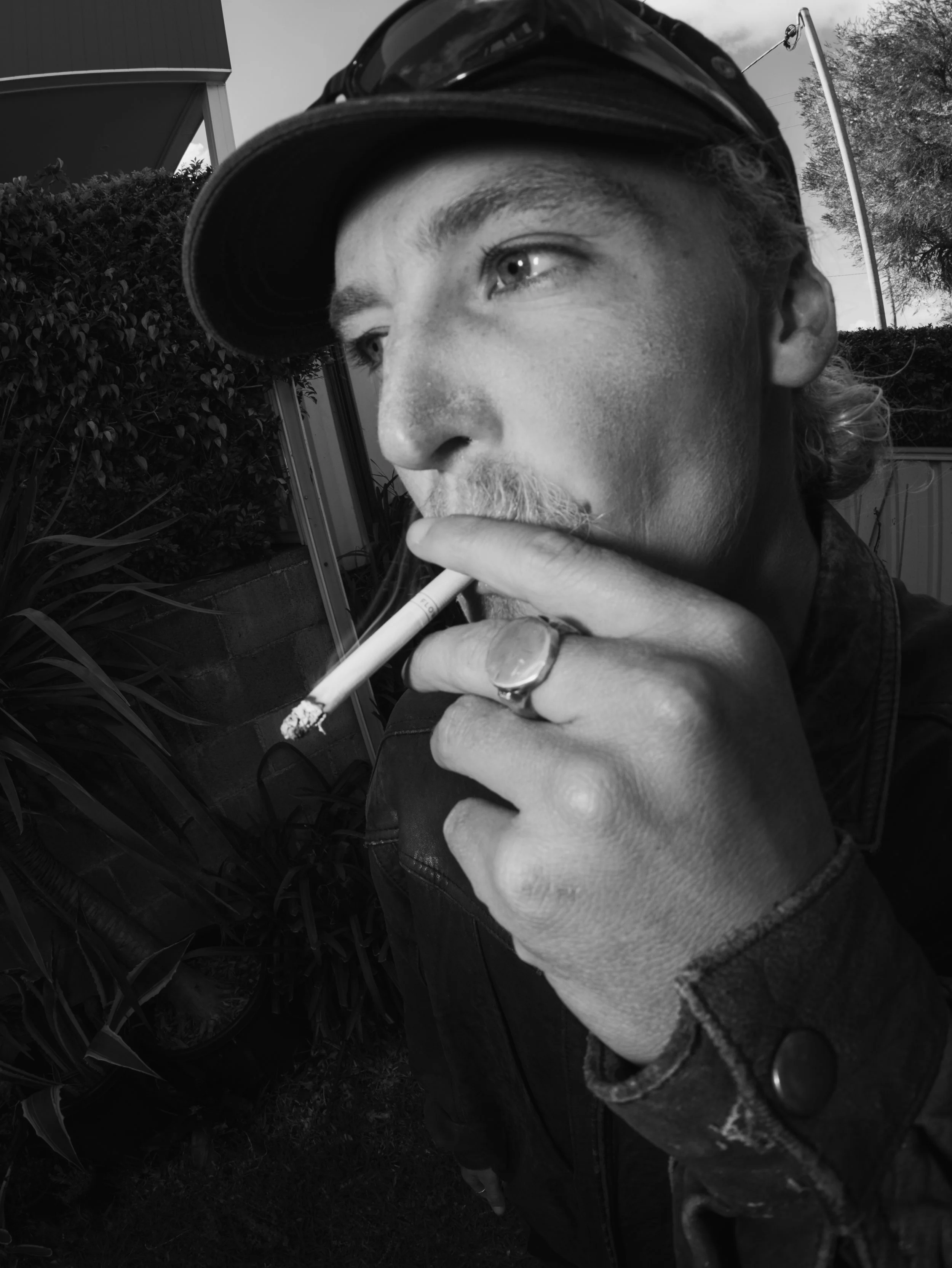 Black-and-white close-up portrait of a bearded man in a baseball cap holding a lit cigarette outdoors