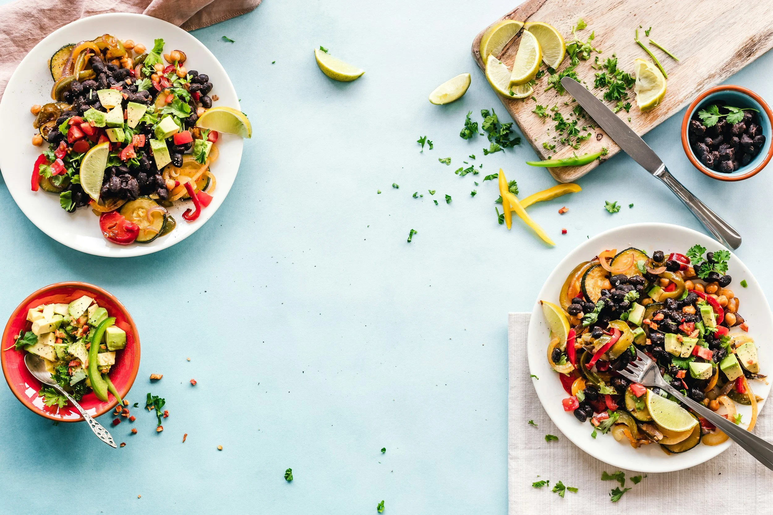 Colorful Mexican-style vegetable salads with black beans, chopped vegetables, avocado, lime wedges, and chopped herbs on white and red bowls and a cutting board on a light blue surface.