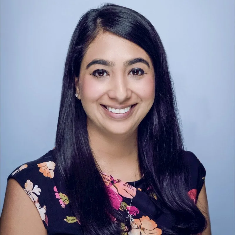 Smiling person with long dark hair, wearing a floral top against a light blue background.
