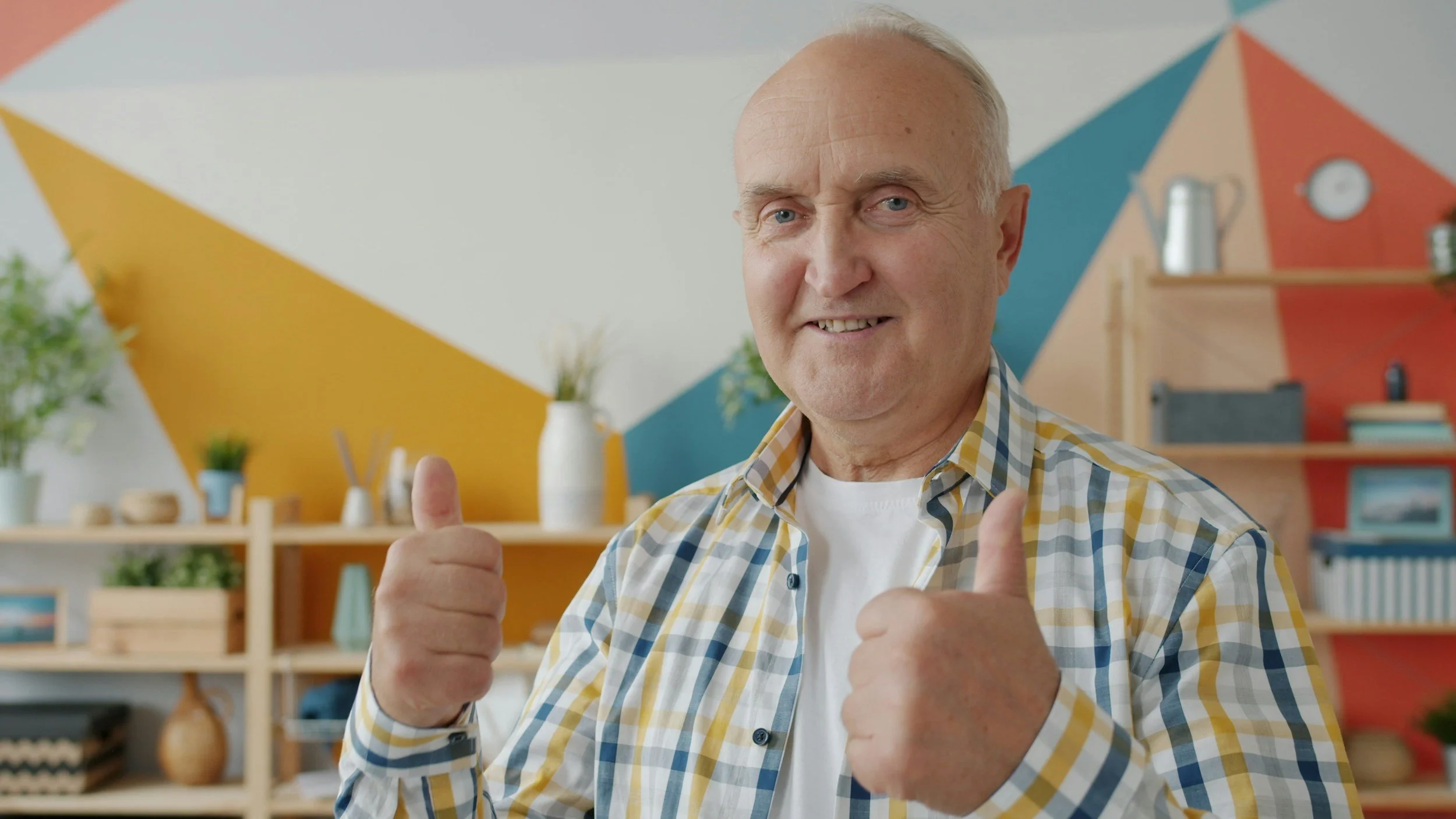 An older man with light skin, dressed in a plaid yellow, blue, and white shirt, smiling and giving a thumbs-up gesture in a colorful room with geometric painted walls, shelves with plants, books, and decorative items.