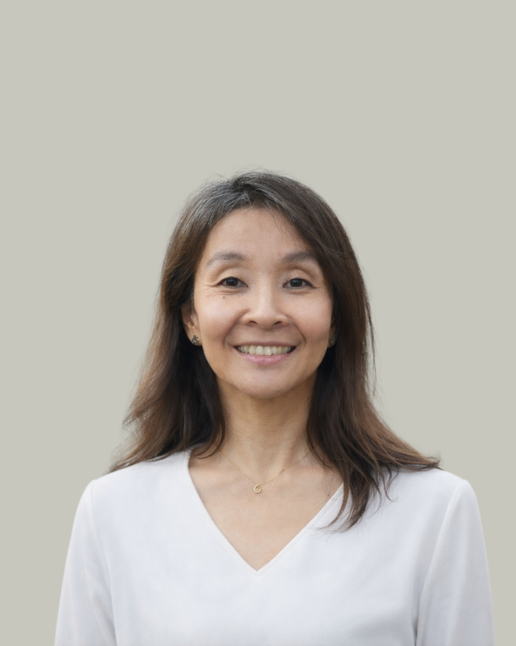 A woman with shoulder-length brown hair, wearing a white V-neck blouse, smiling against a plain, light-colored background.