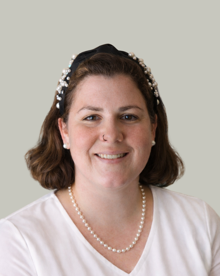A woman with shoulder-length brown hair, wearing a pearl headband, pearl earrings, a pearl necklace, a rose nose ring, and a cream-colored shirt, smiling with a neutral plain background.