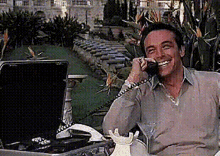 Smiling man on the phone in a garden, beside a table with a laptop.