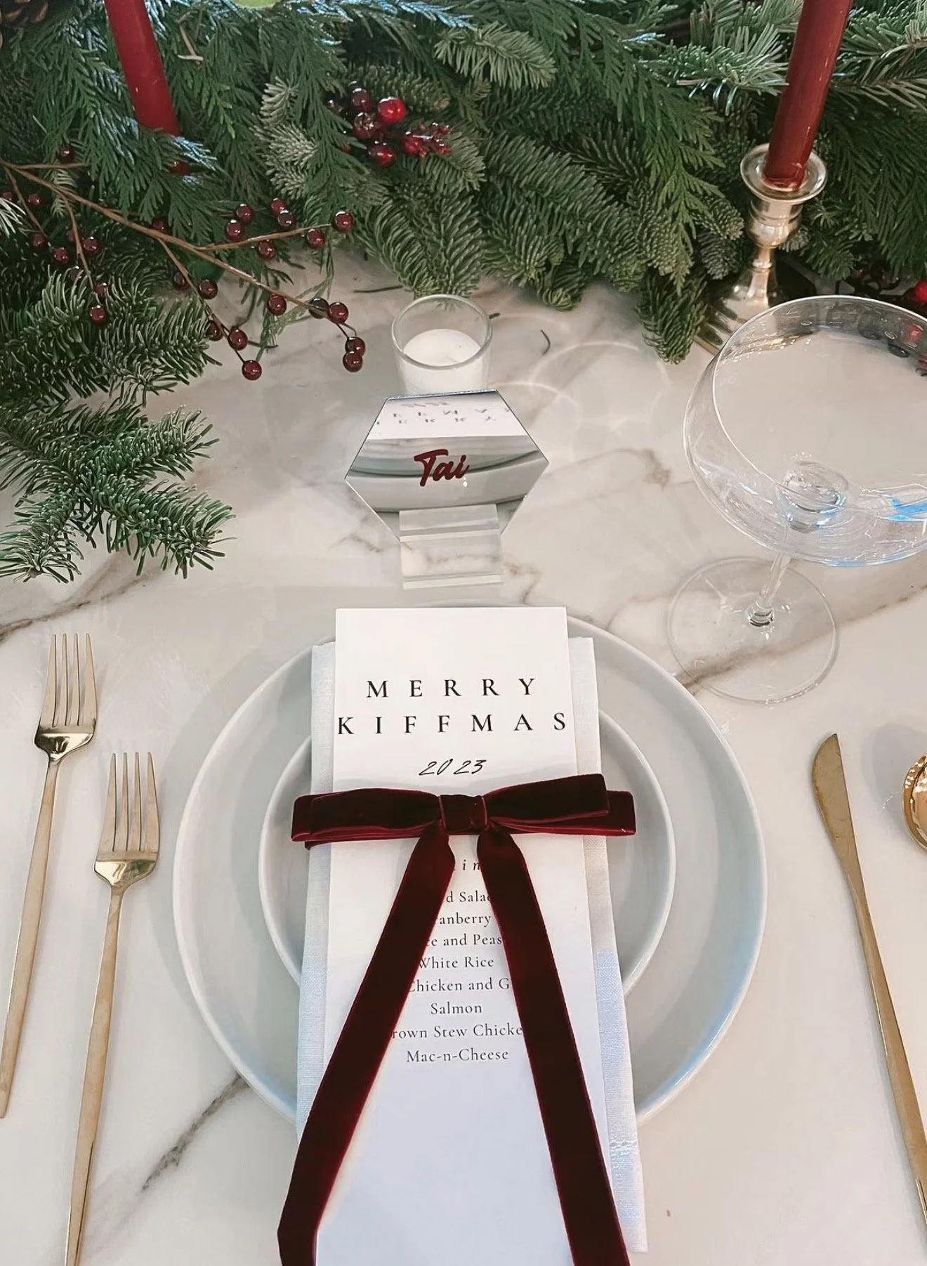 Festive Christmas dinner place setting with a personalized menu card, burgundy velvet ribbon, white plates, gold cutlery, red candles, and evergreen foliage with red berries.