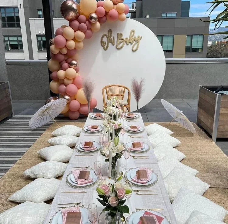 Decorated outdoor dining setup with a long table, white plates, pink napkins, gold cutlery, and floral centerpieces, surrounded by pillows and umbrellas. A balloons arch with pink, cream, and metallic balloons decorates a white circular backdrop with