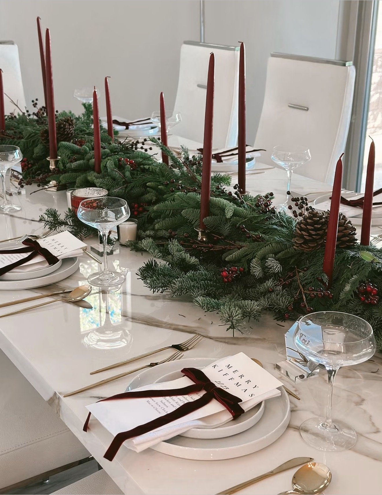 Elegant Christmas dinner table decorated with evergreen garlands, burgundy taper candles, red berries, white plates, gold cutlery, crystal glassware, and burgundy ribbon accents.