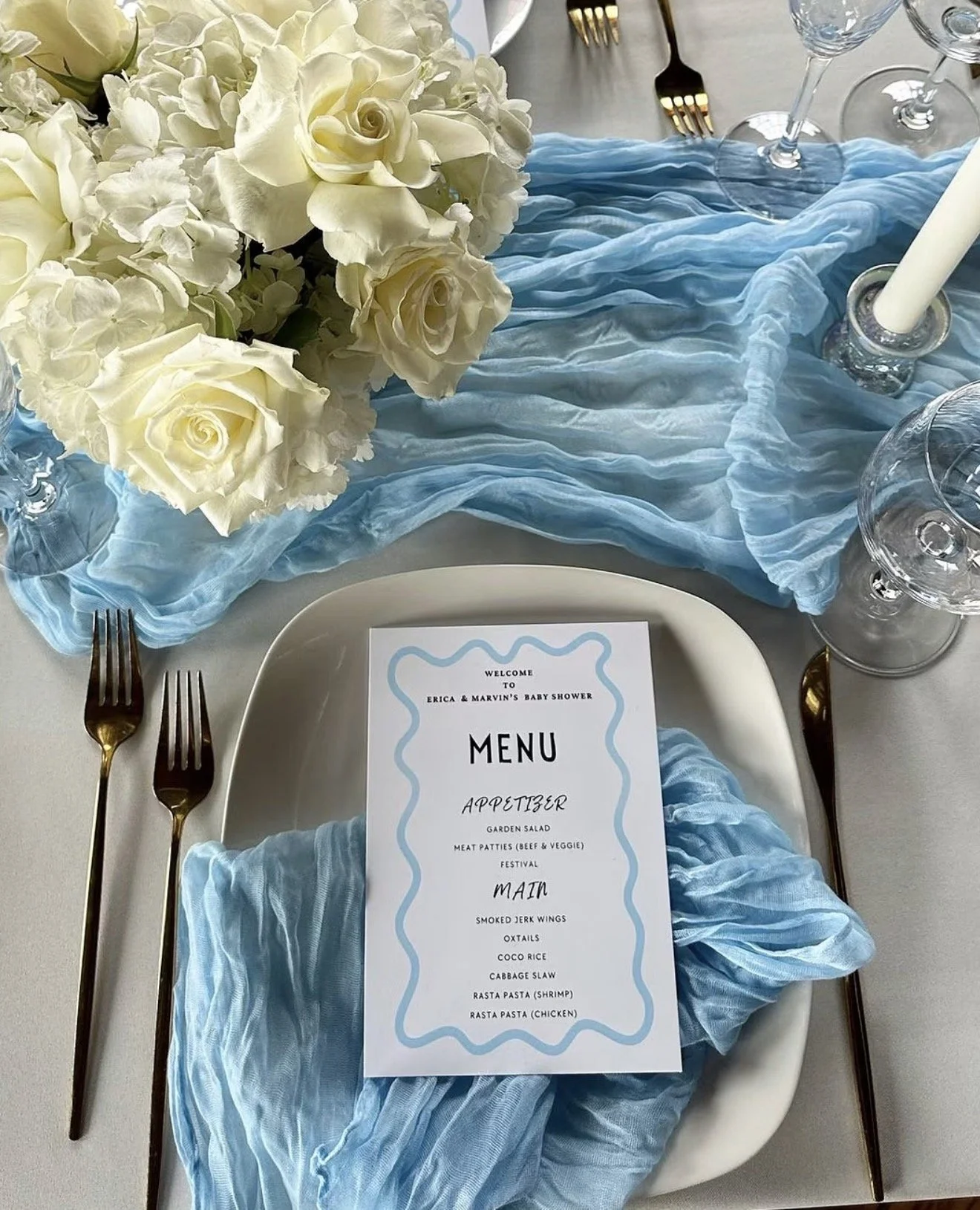 Elegant baby shower table setting featuring white roses, flowing blue fabric, gold cutlery, crystal glassware, and a personalized menu card on a white plate.