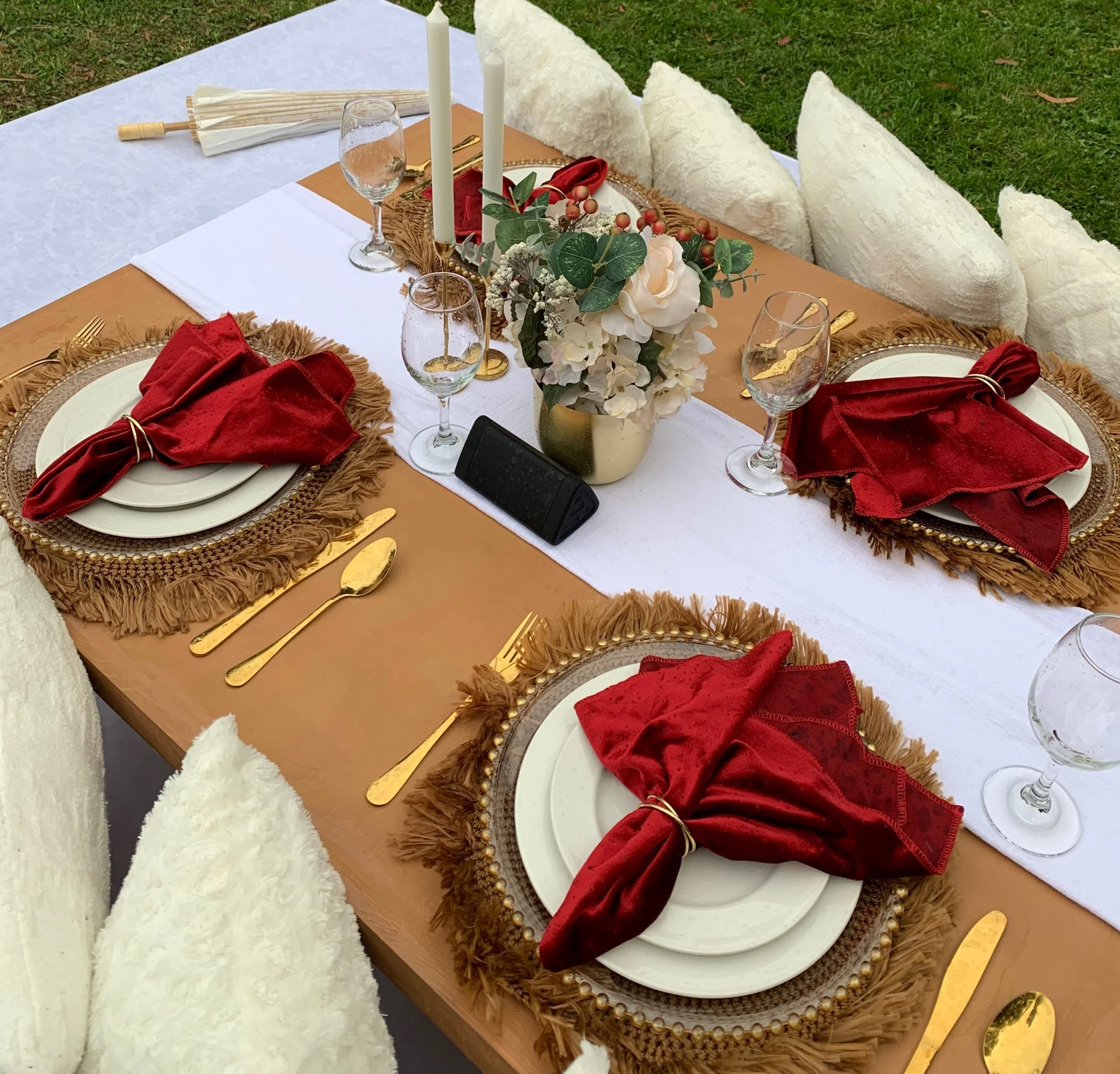 Elegant outdoor Friendsgiving picnic table featuring white plates, gold cutlery, red cloth napkins, fringed placemats, floral arrangements, and plush white cushions on a grassy lawn.