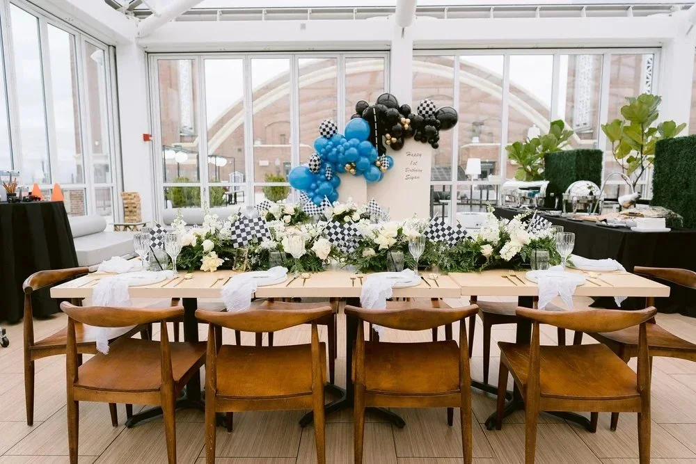 A long dining table decorated with white and green floral arrangements, checkered black and white flags, and place settings with white napkins, glasses, and utensils. Behind the table, there is a balloon arch with black, blue, and checkered balloons,