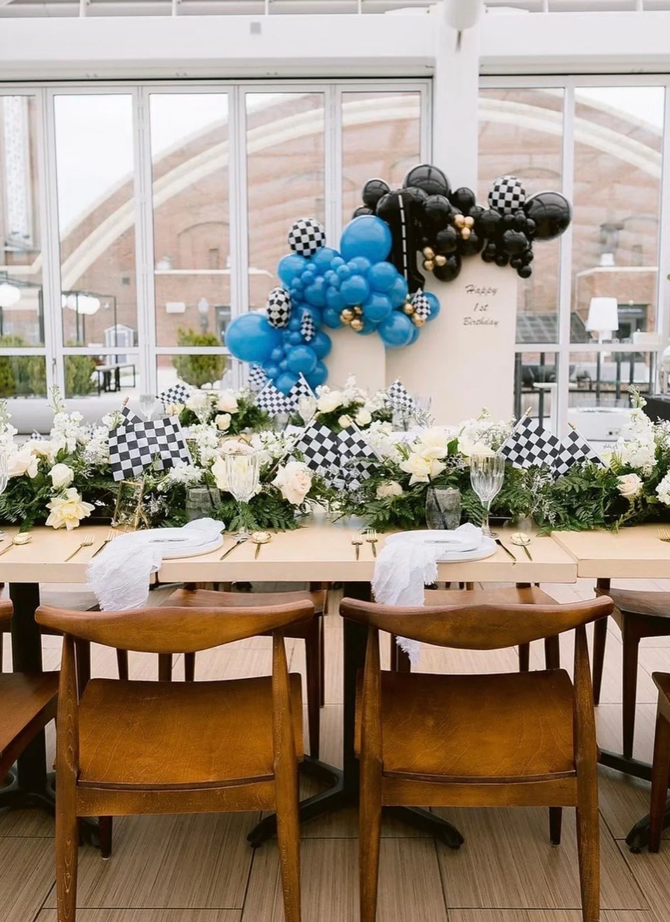 Race car themed birthday party tablescape featuring blue and black balloons, checkered flags, white flowers, and elegant wooden chairs.