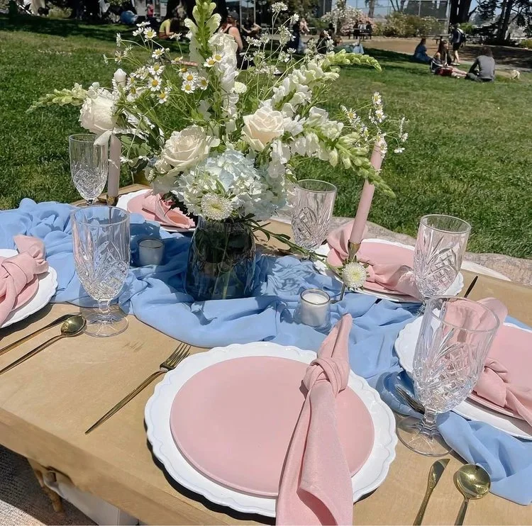 A table set for an outdoor event with pink paper plates, pink cloth napkins, clear crystal glasses, gold utensils, and a large floral centerpiece with white roses, daisies, and other white flowers. The table has a blue fabric runner, and small candle