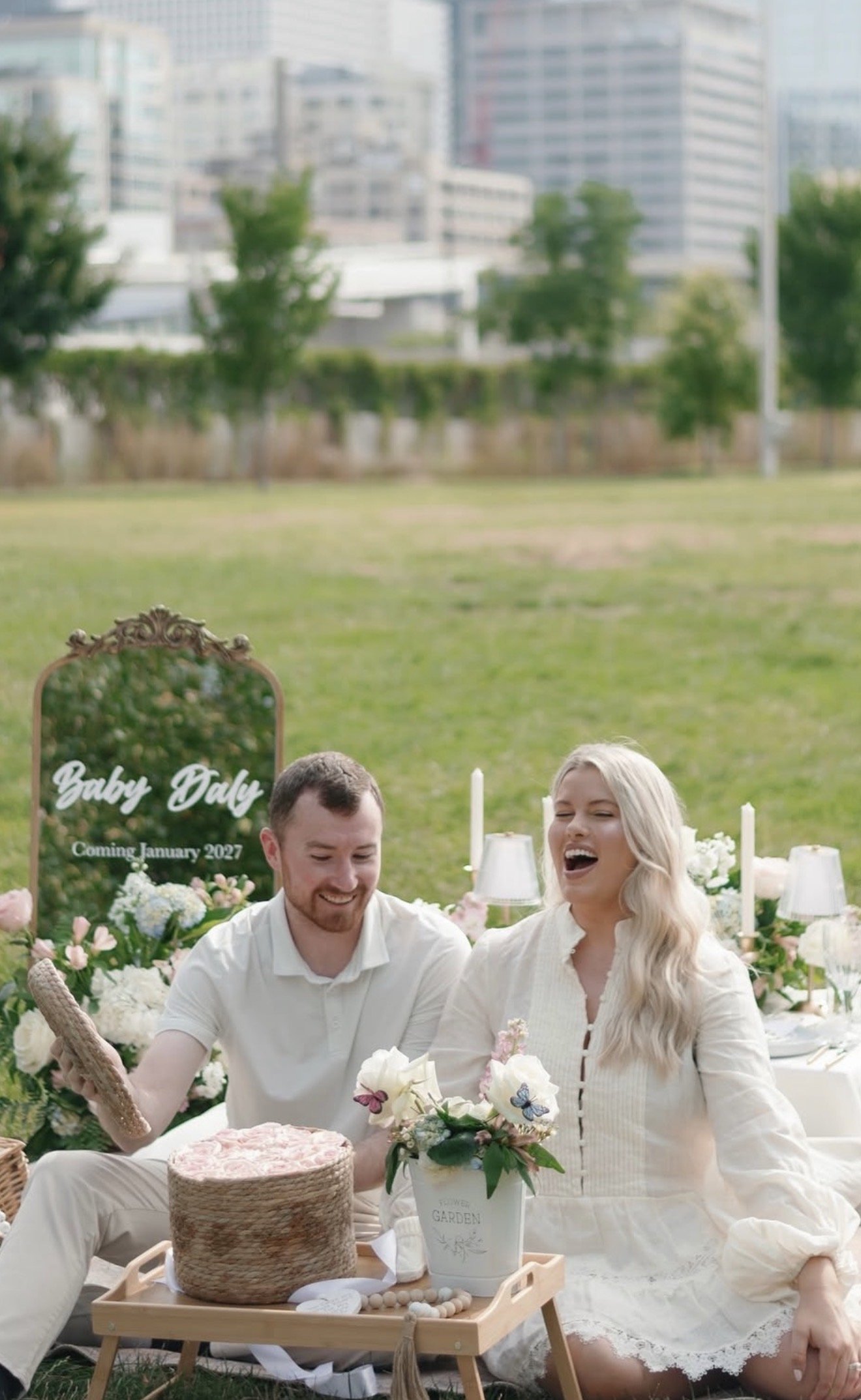 Couple celebrating an outdoor baby shower at a picnic table decorated with pastel flowers, woven baskets, white cushions, and a sign announcing the baby's expected arrival in January 2027.
