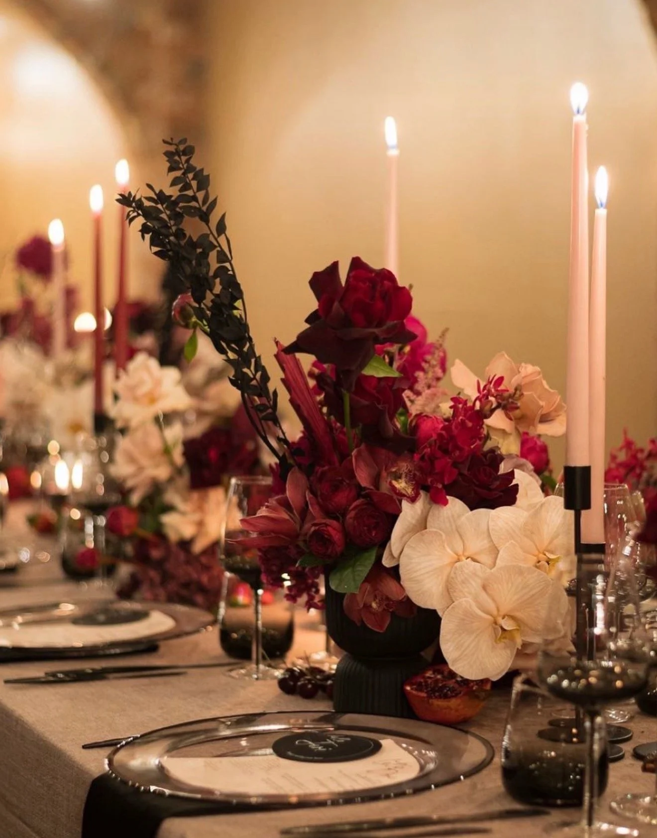 Elegant dinner tablescape featuring burgundy and ivory floral arrangements, tall taper candles, crystal glassware, and sophisticated place settings in warm candlelight.