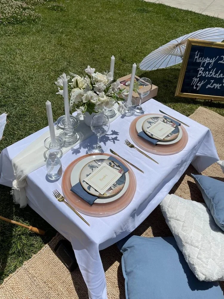 A beautifully set outdoor birthday dinner table with white tablecloth, floral centerpiece, pink and white plates, gold utensils, wine glasses, white candles, a birthday card, and a sign with birthday message, decorated with an umbrella and cushions.