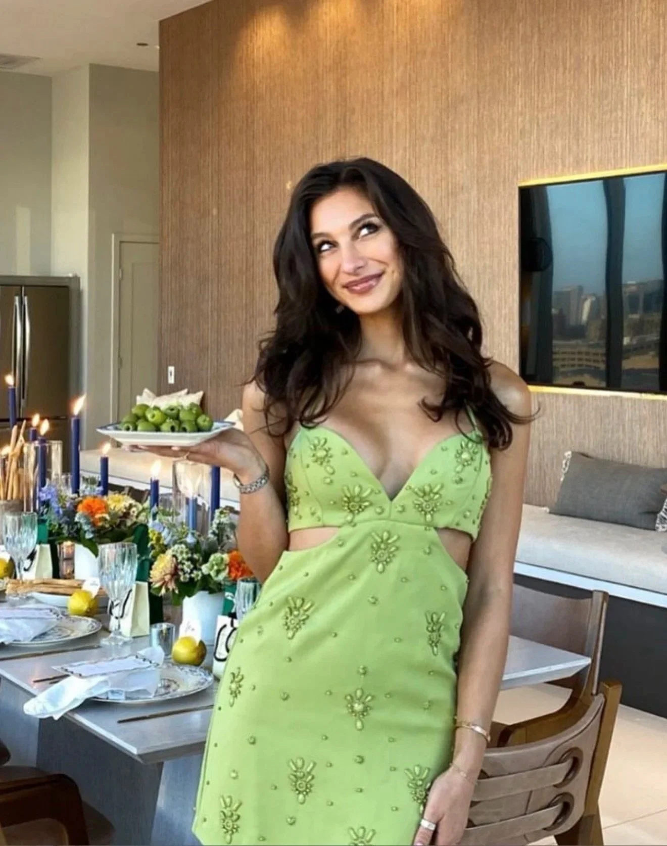 Woman in a green dress posing beside an elegant dining table styled with fresh flowers, decorative glassware, and premium table settings.