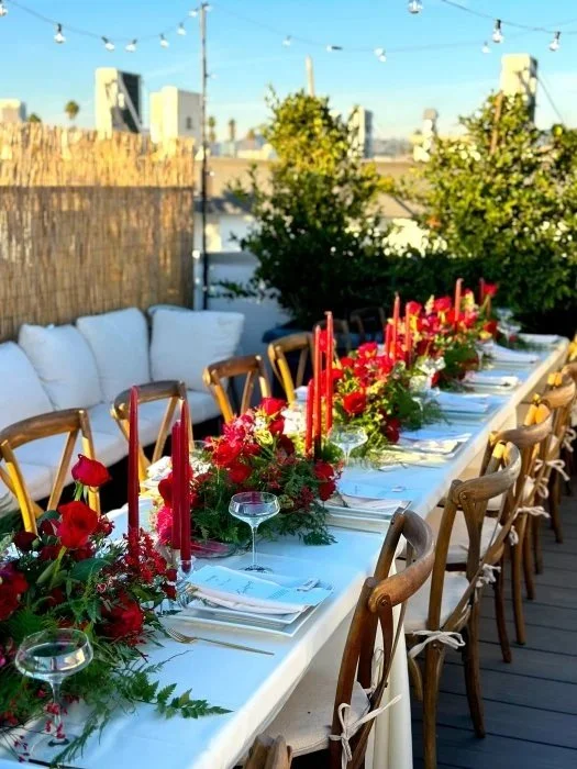Outdoor dining table decorated with red candles and floral arrangements, set with wine glasses and plates on a rooftop or patio with string lights and greenery.